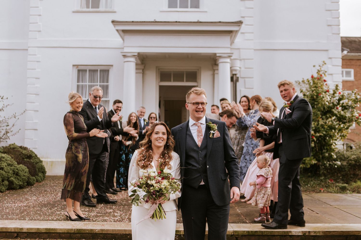 Wedding party members line up outside a white building entrance during a formal celebration.