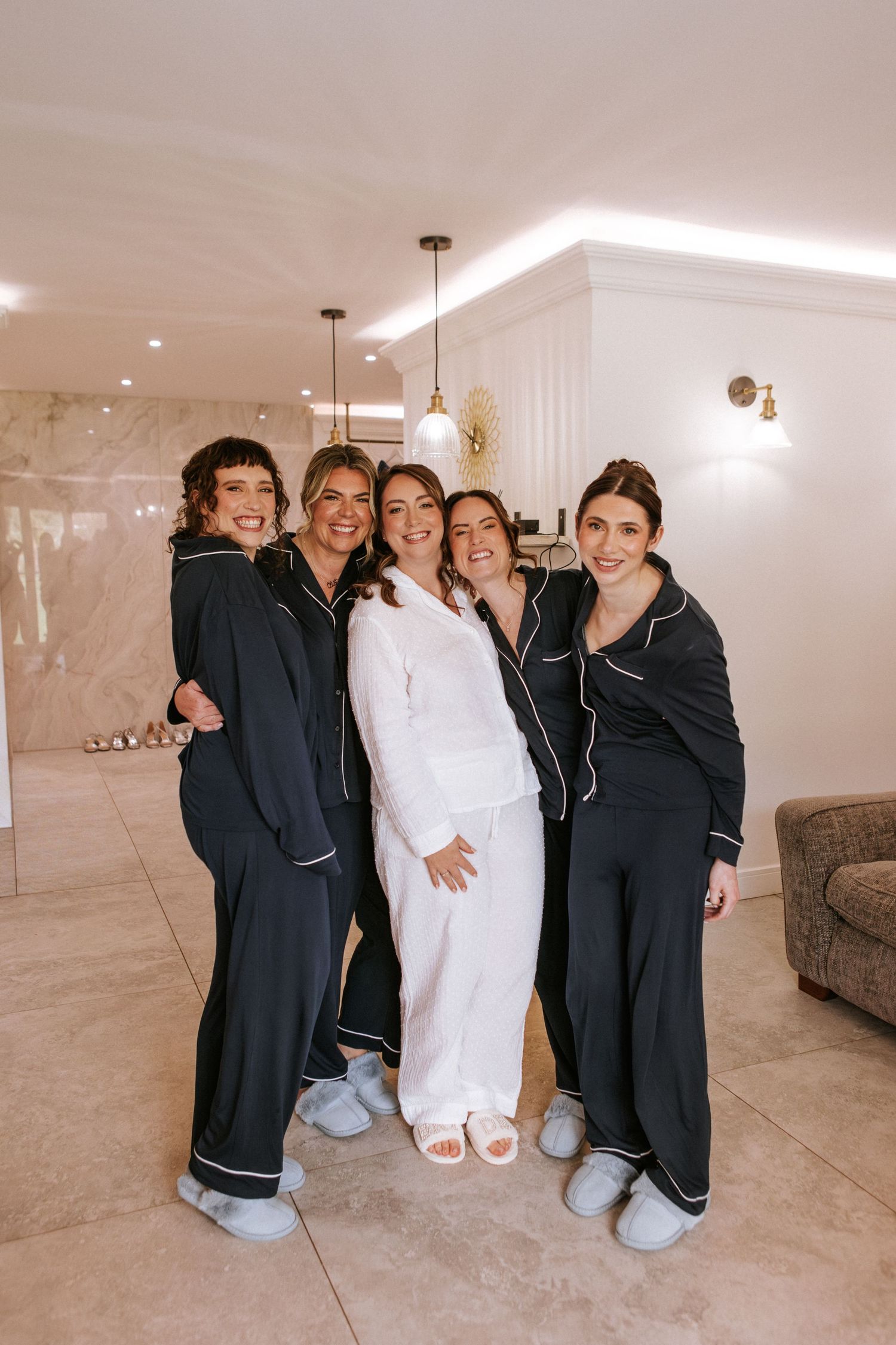 Bride in white satin pajamas poses with four bridesmaids in matching black satin pajamas in an elegant hotel suite.