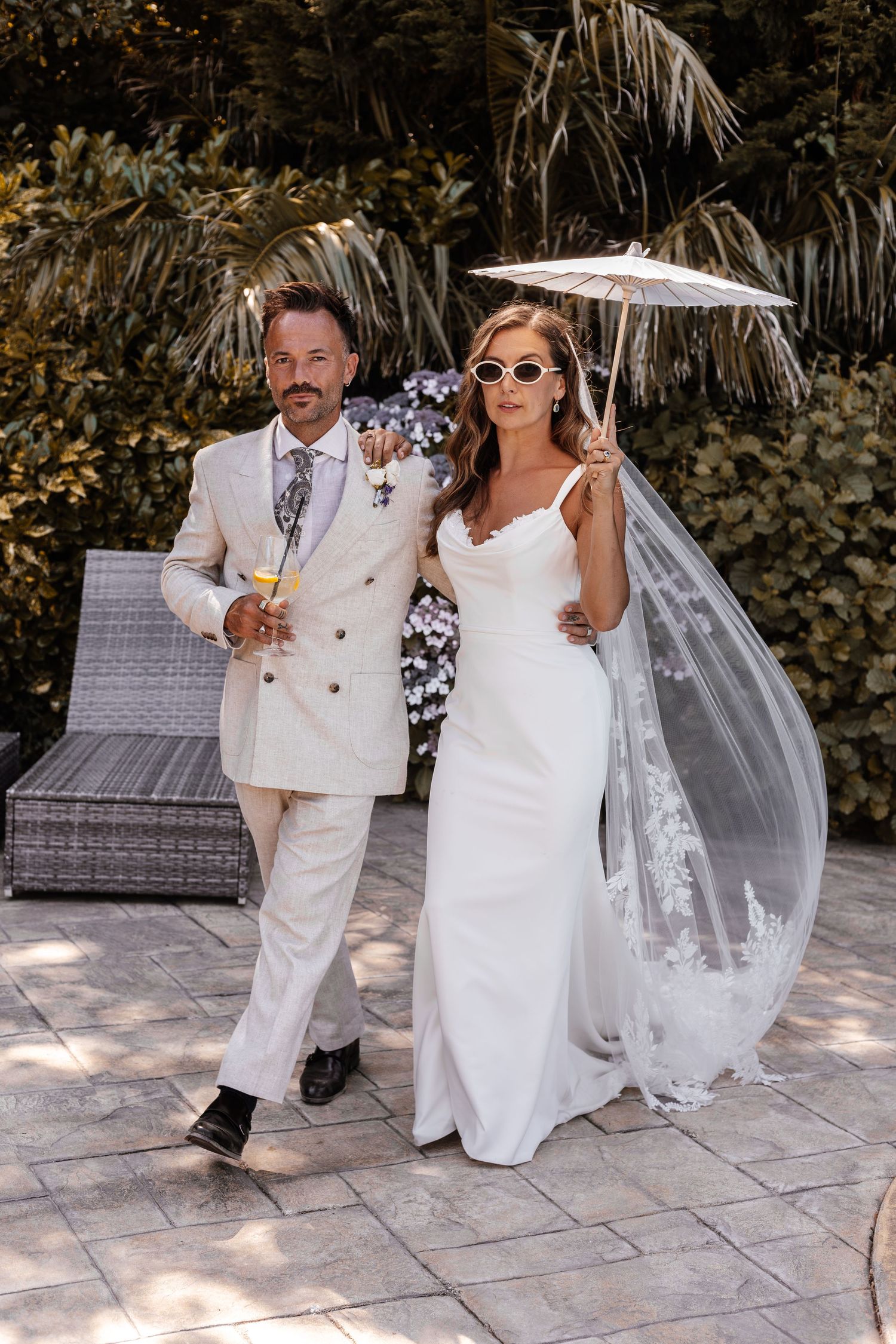 Bride in white gown with veil and sunglasses holds parasol beside groom in cream suit outdoors.