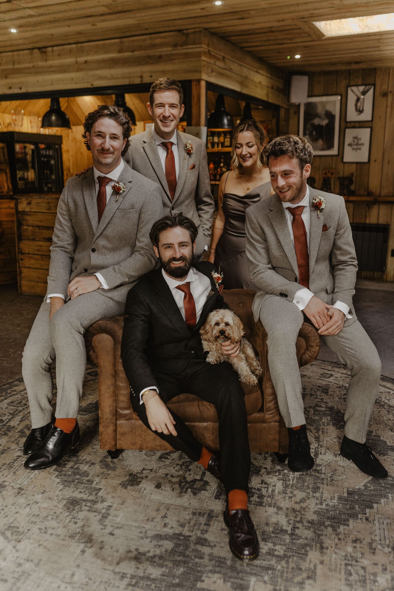 Groomsmen in light gray suits with red ties pose with a small dog in a rustic wooden venue.
