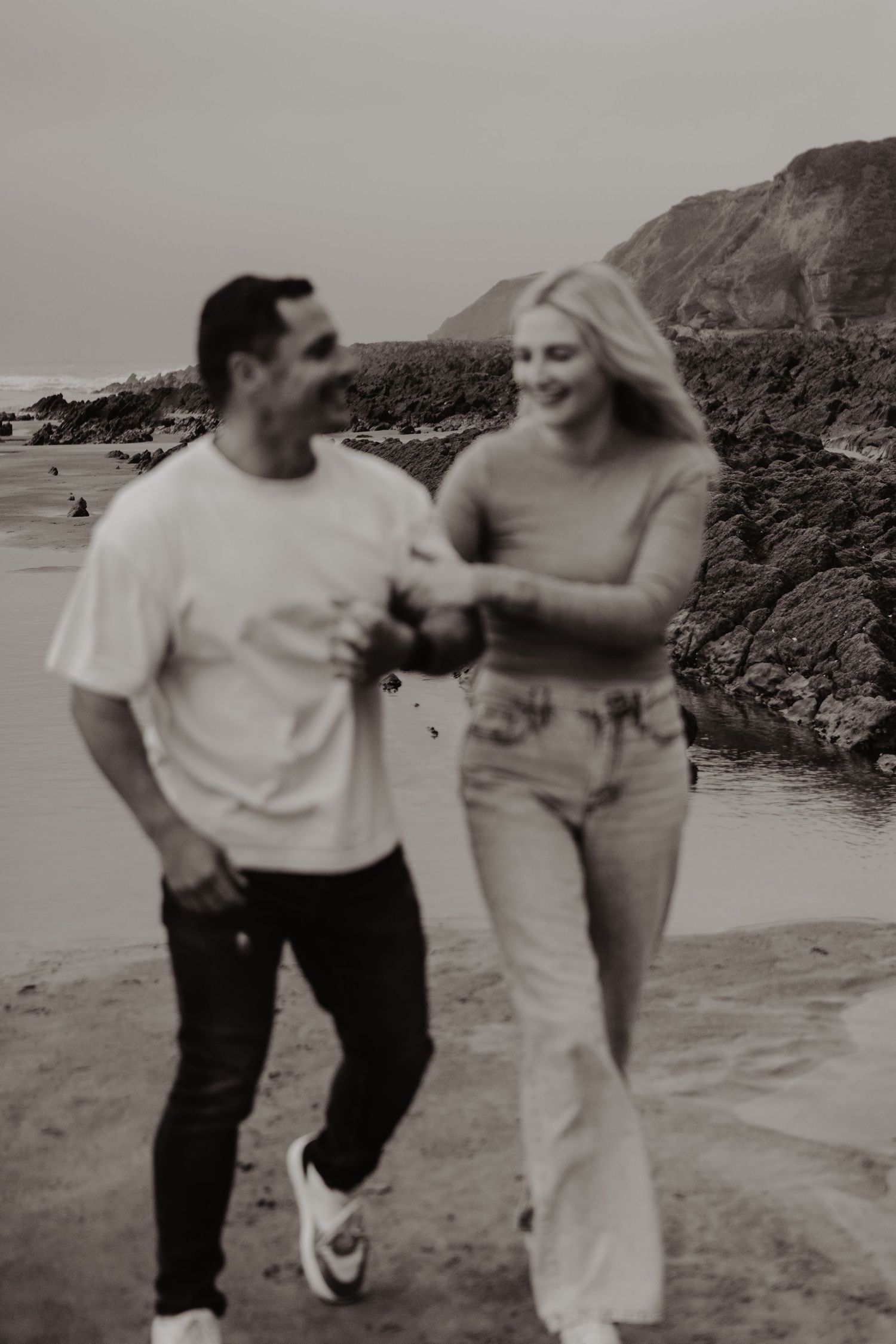Black and white photo of a playful couple dancing on a beach near rocky cliffs.