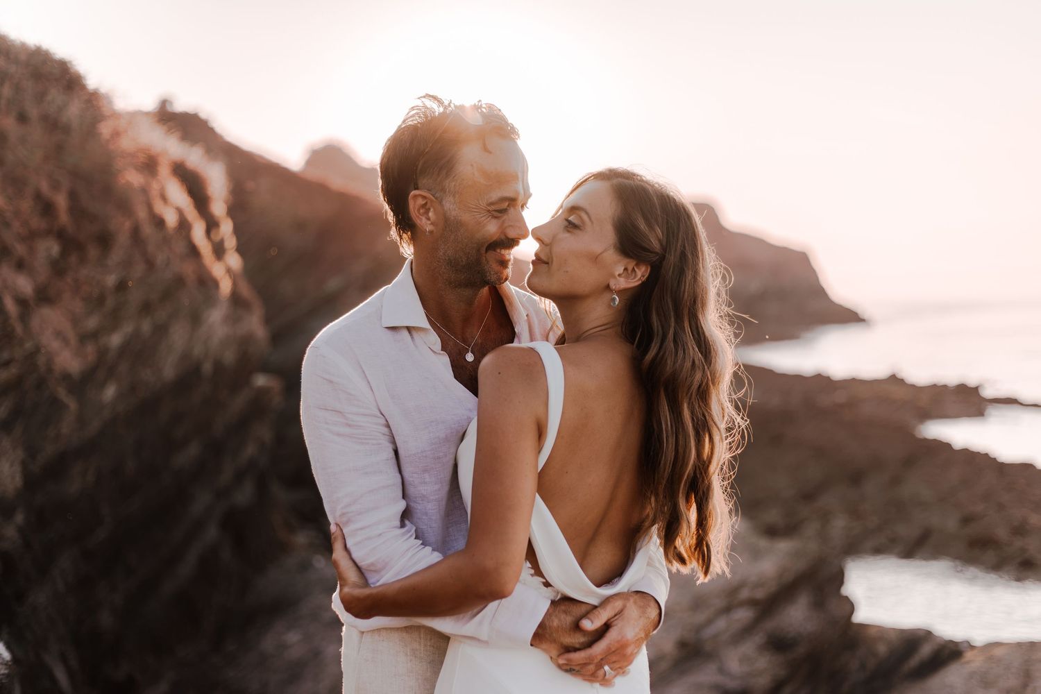 Couple embracing and smiling at sunset on rocky coastal cliffs, golden hour engagement photo.