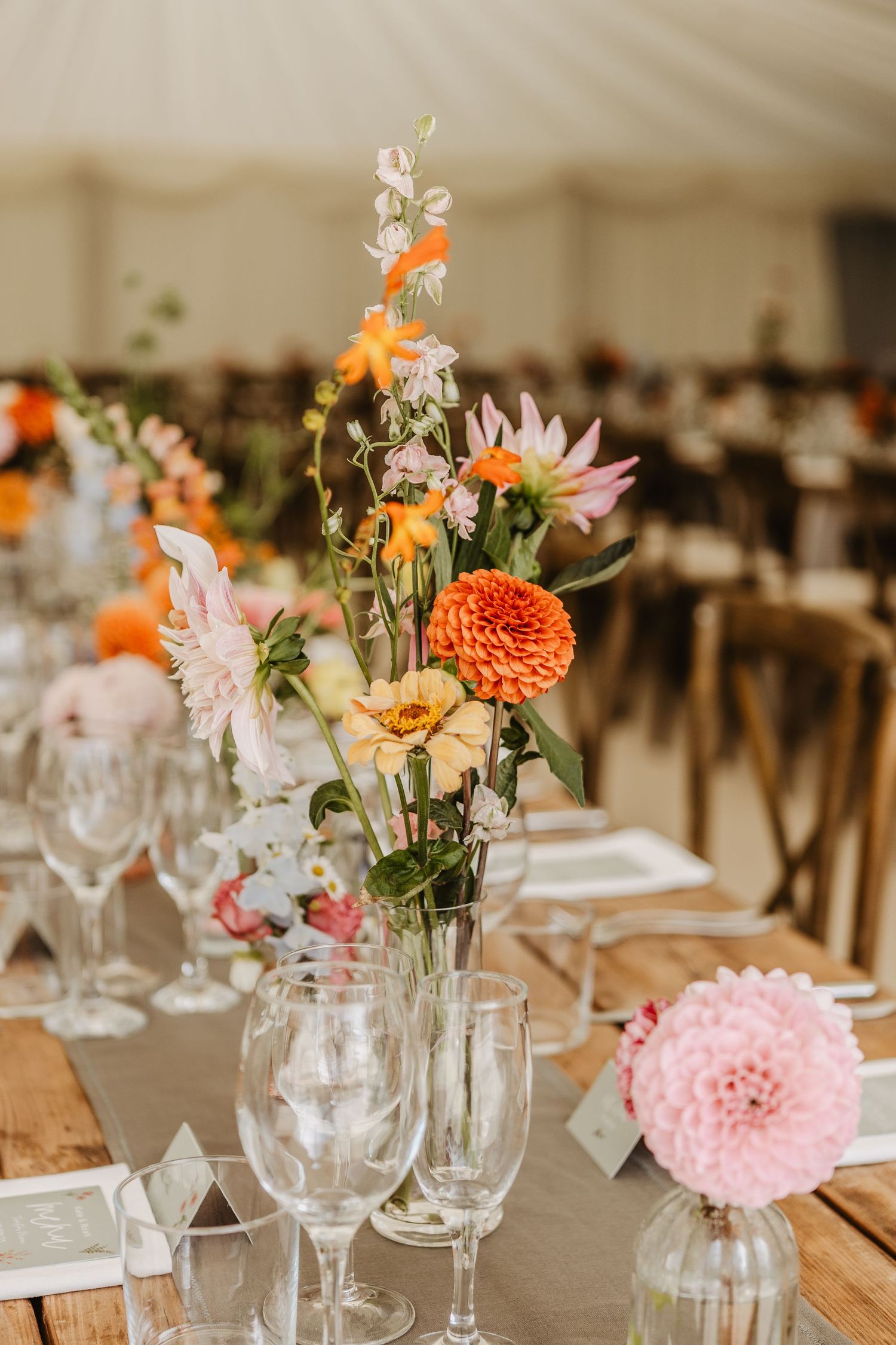 Wedding reception table settings with mason jar flower centerpieces featuring pink and orange dahlias on a natural linen tablecloth.