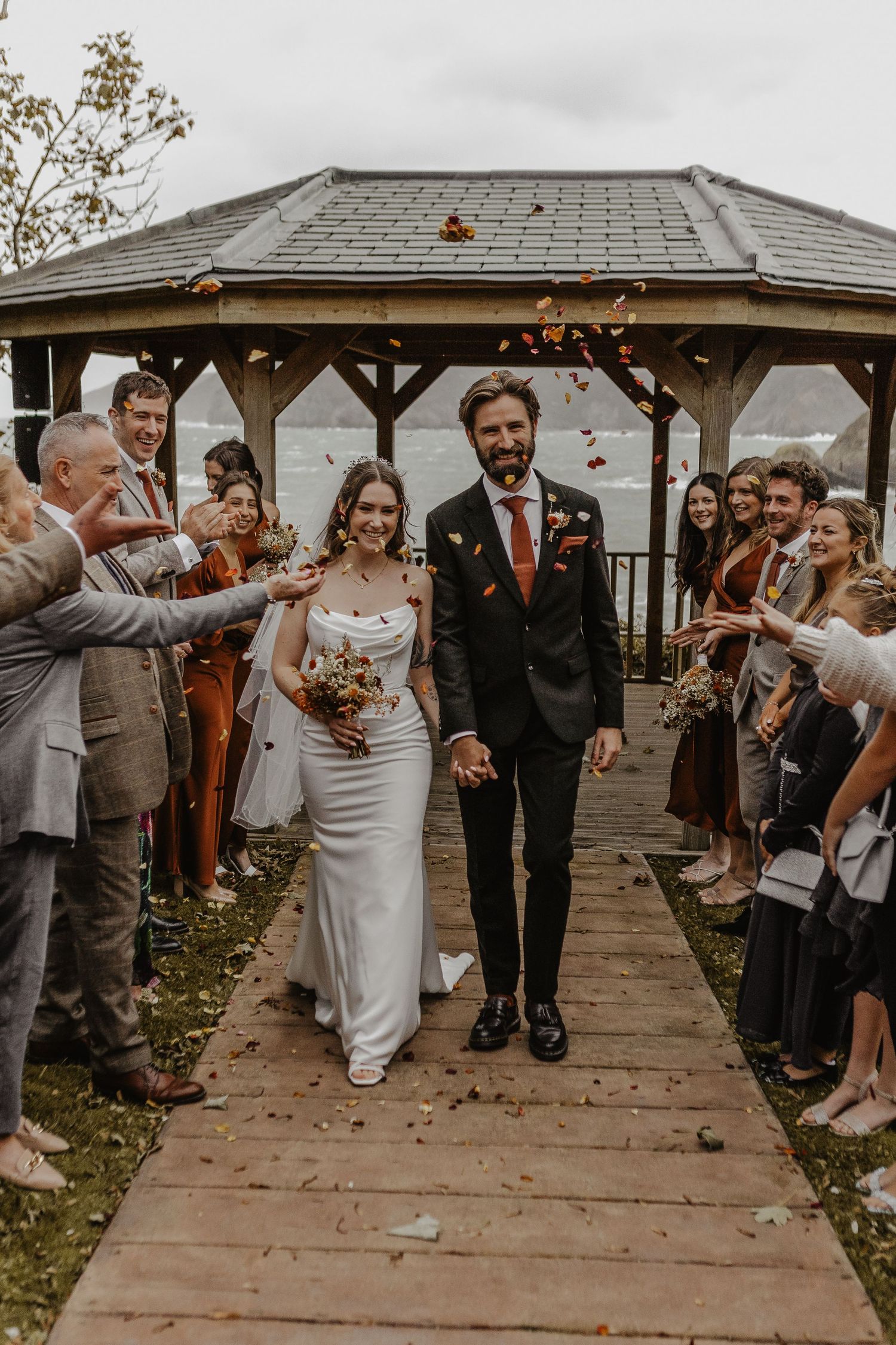 Guests throw petals as newlyweds exit under wooden gazebo.