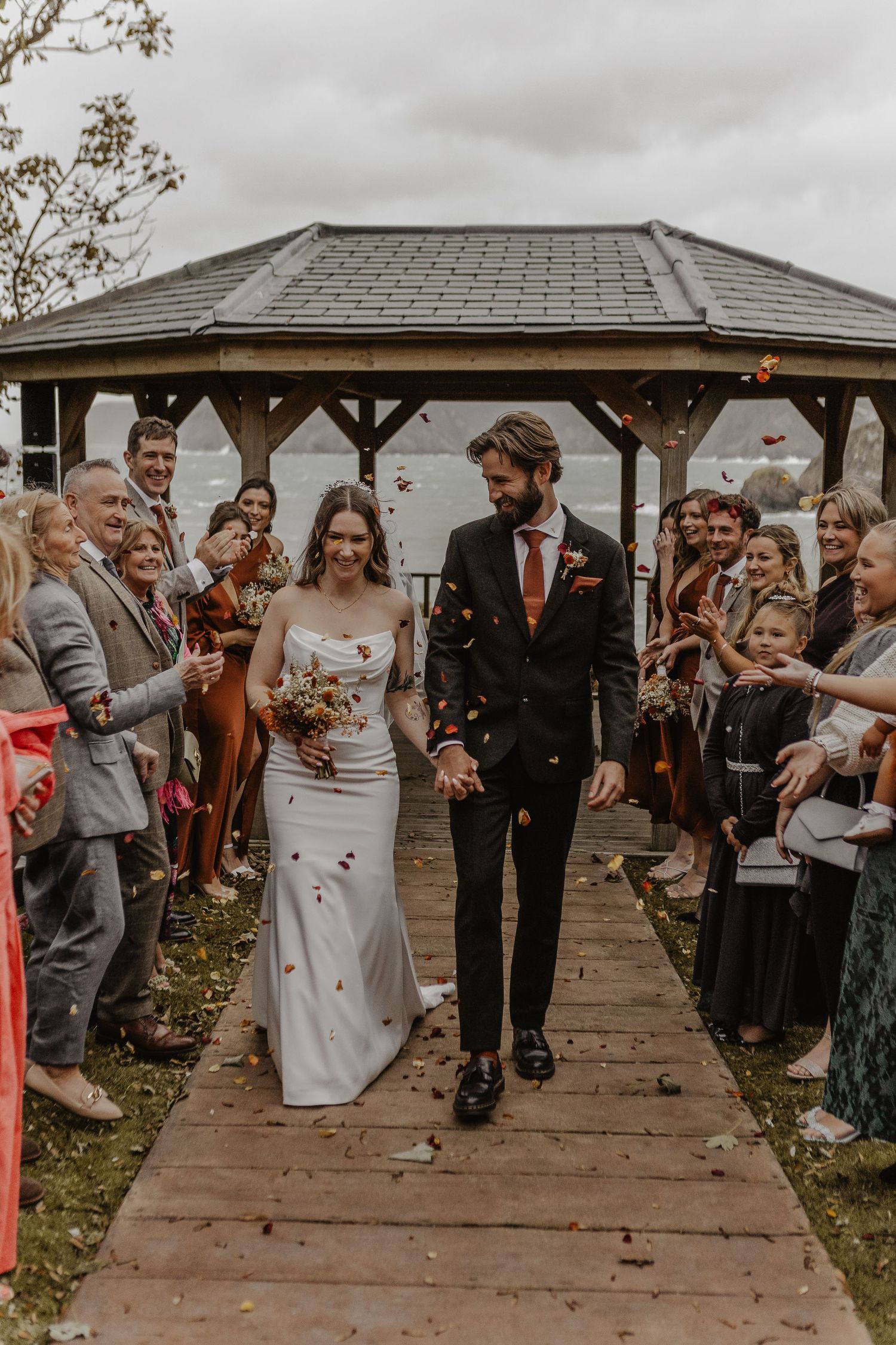 Wedding guests celebrate by throwing confetti as newlyweds walk under a covered outdoor pavilion.