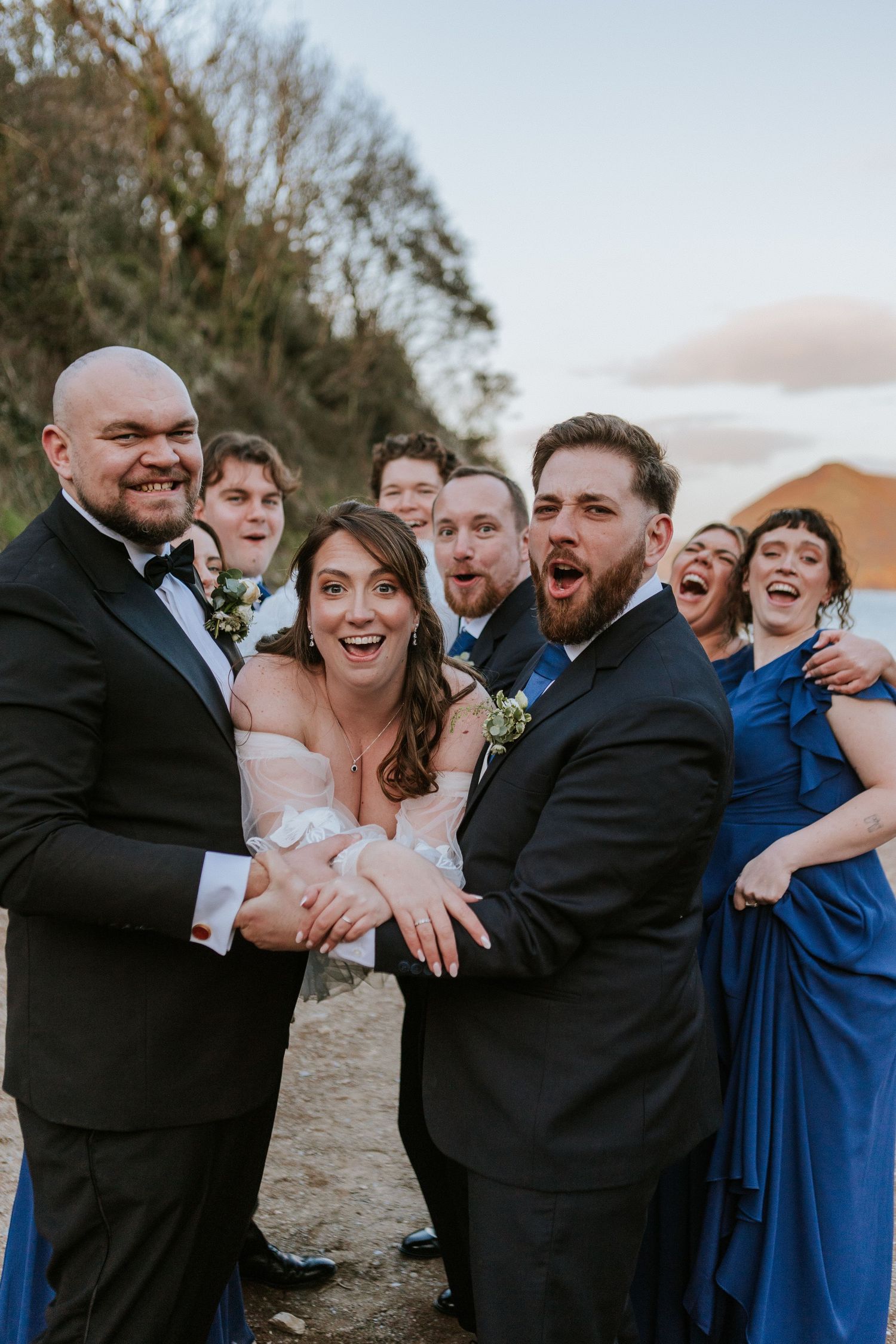 Wedding party in blue and black attire laughing and showing off rings outdoors at golden hour.