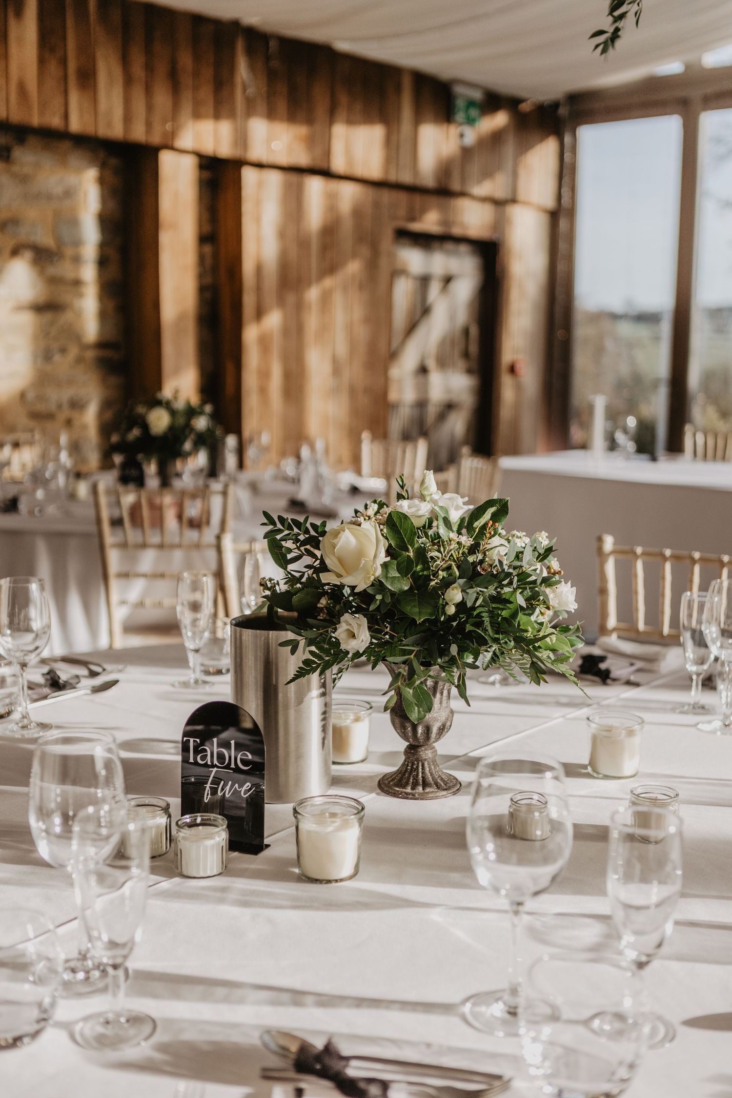 Elegant wedding reception tables with white linens, greenery centerpieces and rustic wooden beams in a barn venue.