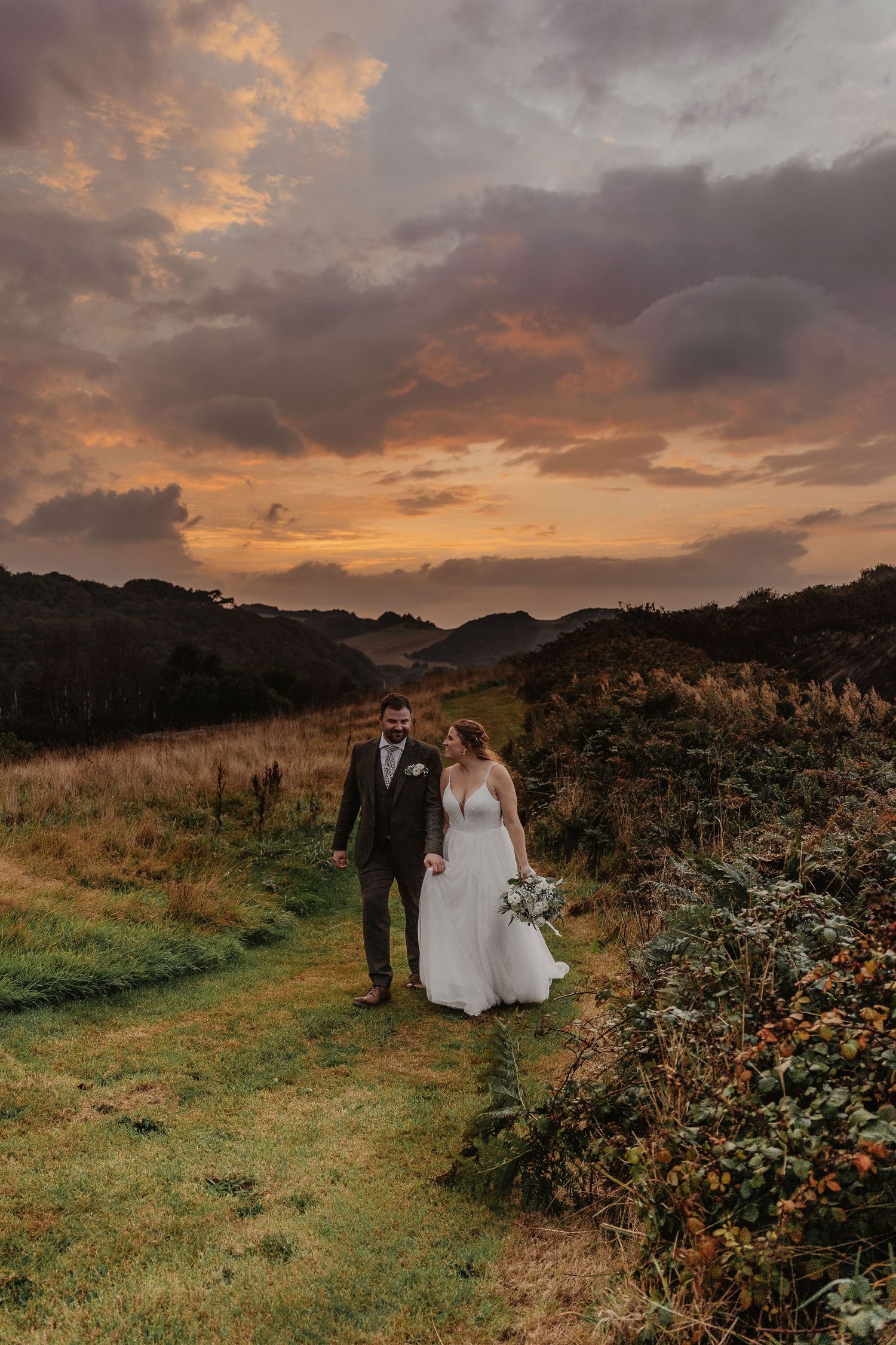 A couple walks along a coastal path through wild blackberry bushes at sunset with dramatic orange clouds overhead.