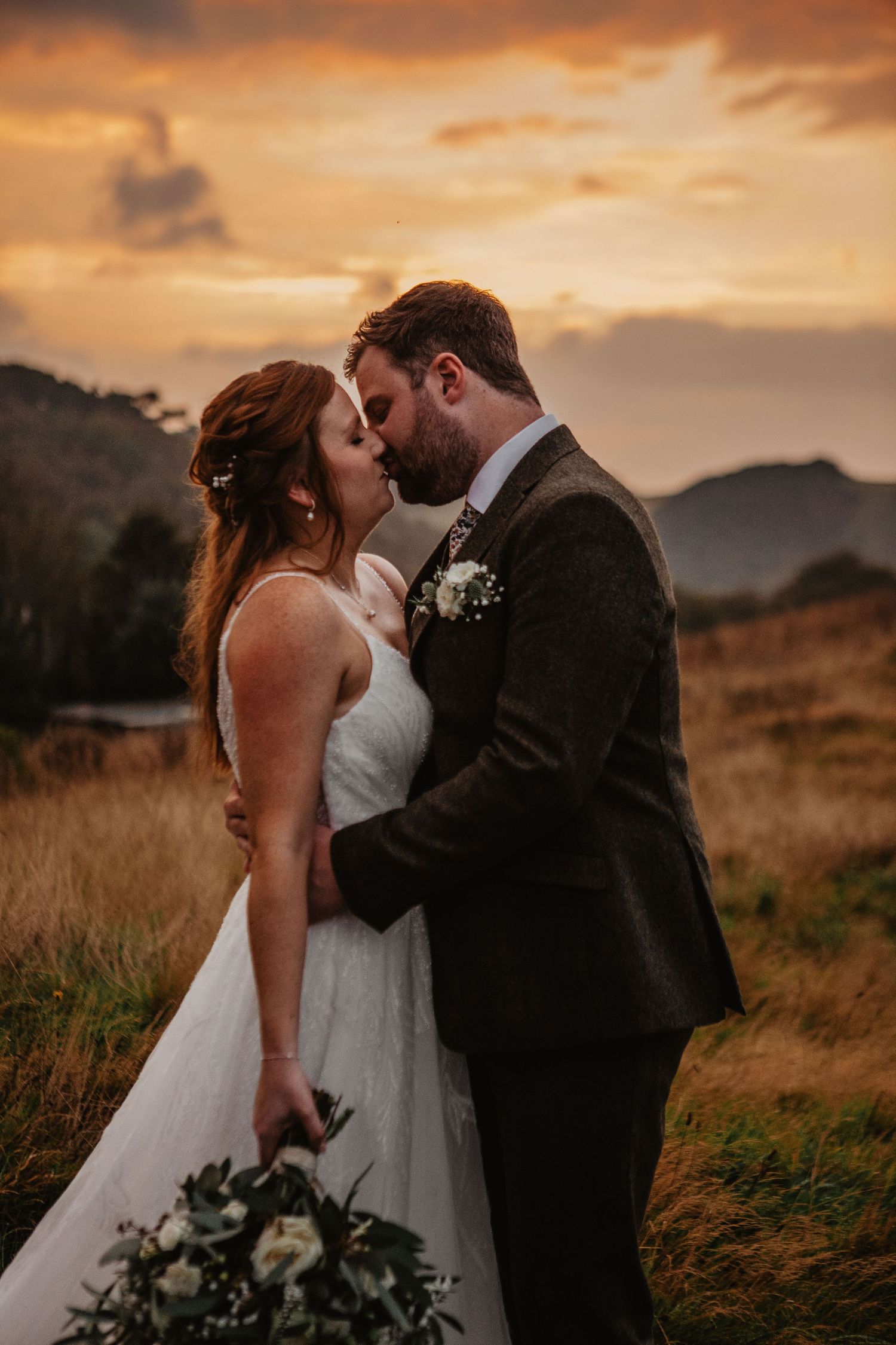 A romantic wedding couple embraces during golden sunset in a mountainous field with tall grass and dramatic sky.