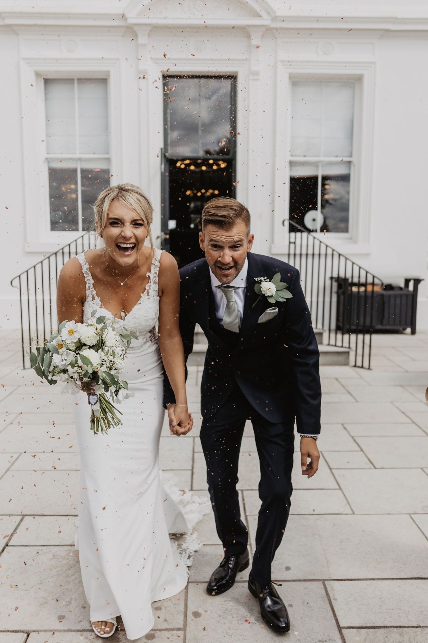 Joyful couple laughing together on a white building exterior during their wedding celebration.