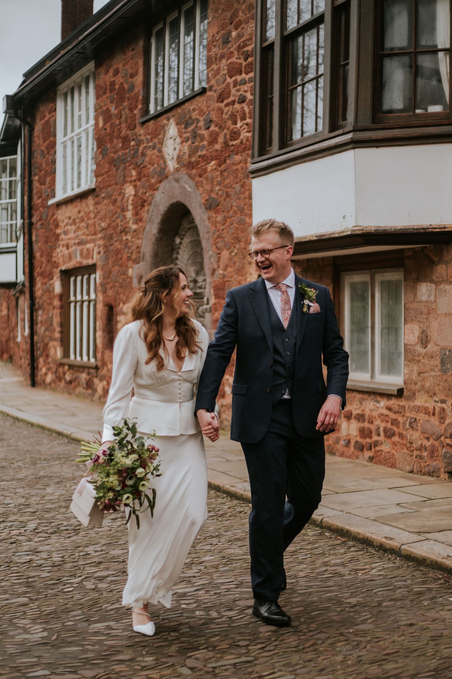 A couple in wedding attire walking along a cobblestone street beside a rustic brick building.