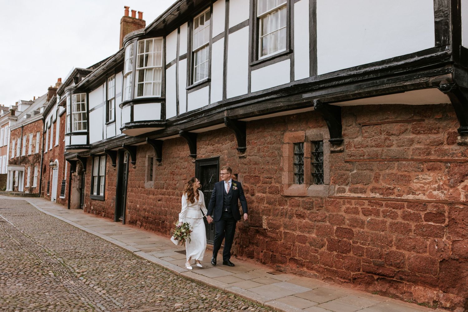 A couple walks down a charming cobblestone street lined with historic red brick buildings.