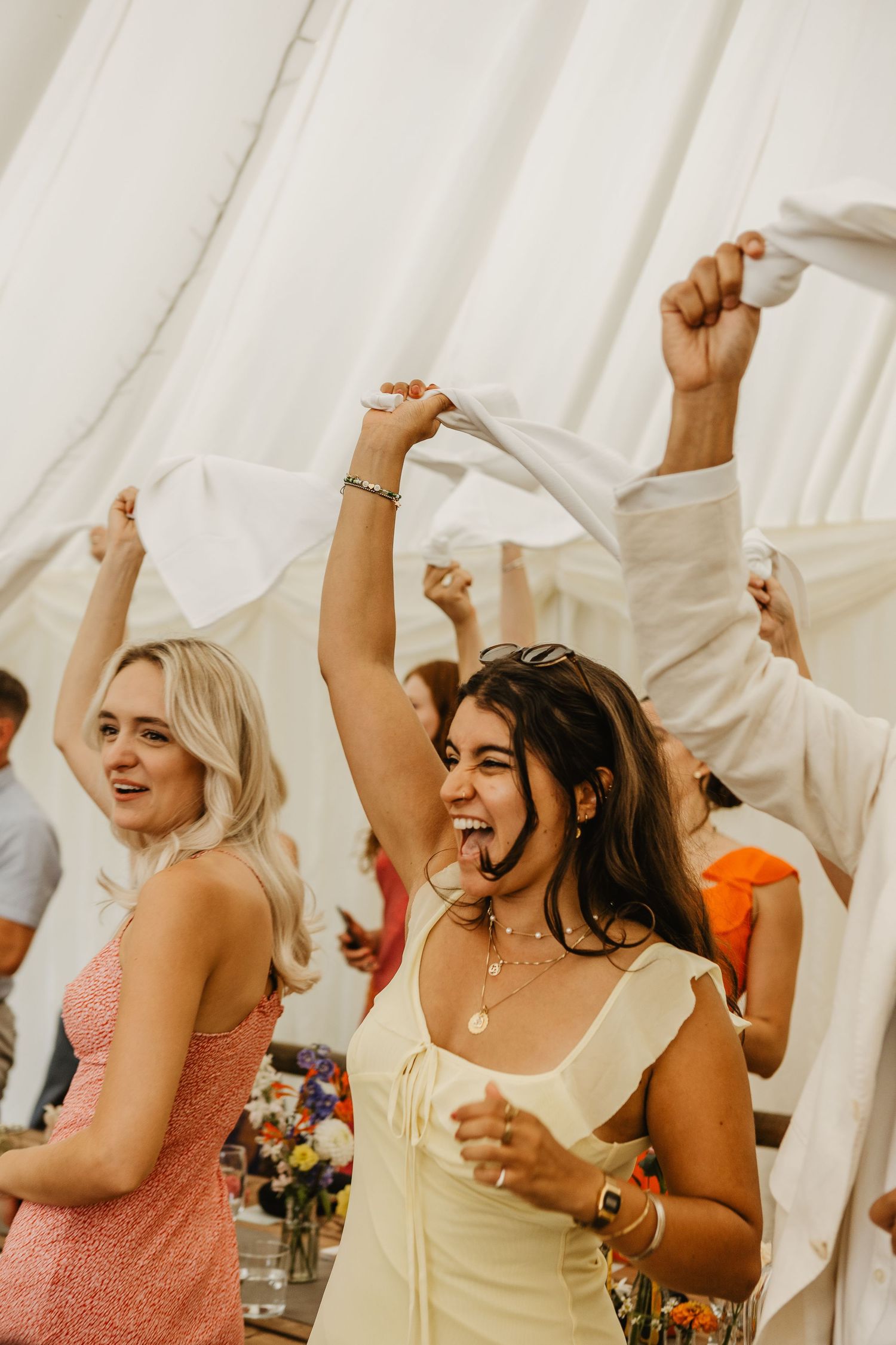 Wedding guests dance and celebrate with napkins in the air at a joyful reception under a white tent.