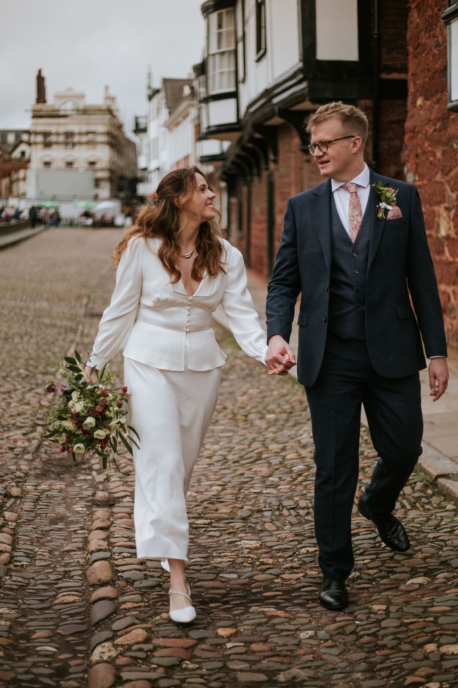 Couple walking together on cobblestone street in wedding attire with bride in white dress and groom in dark suit.