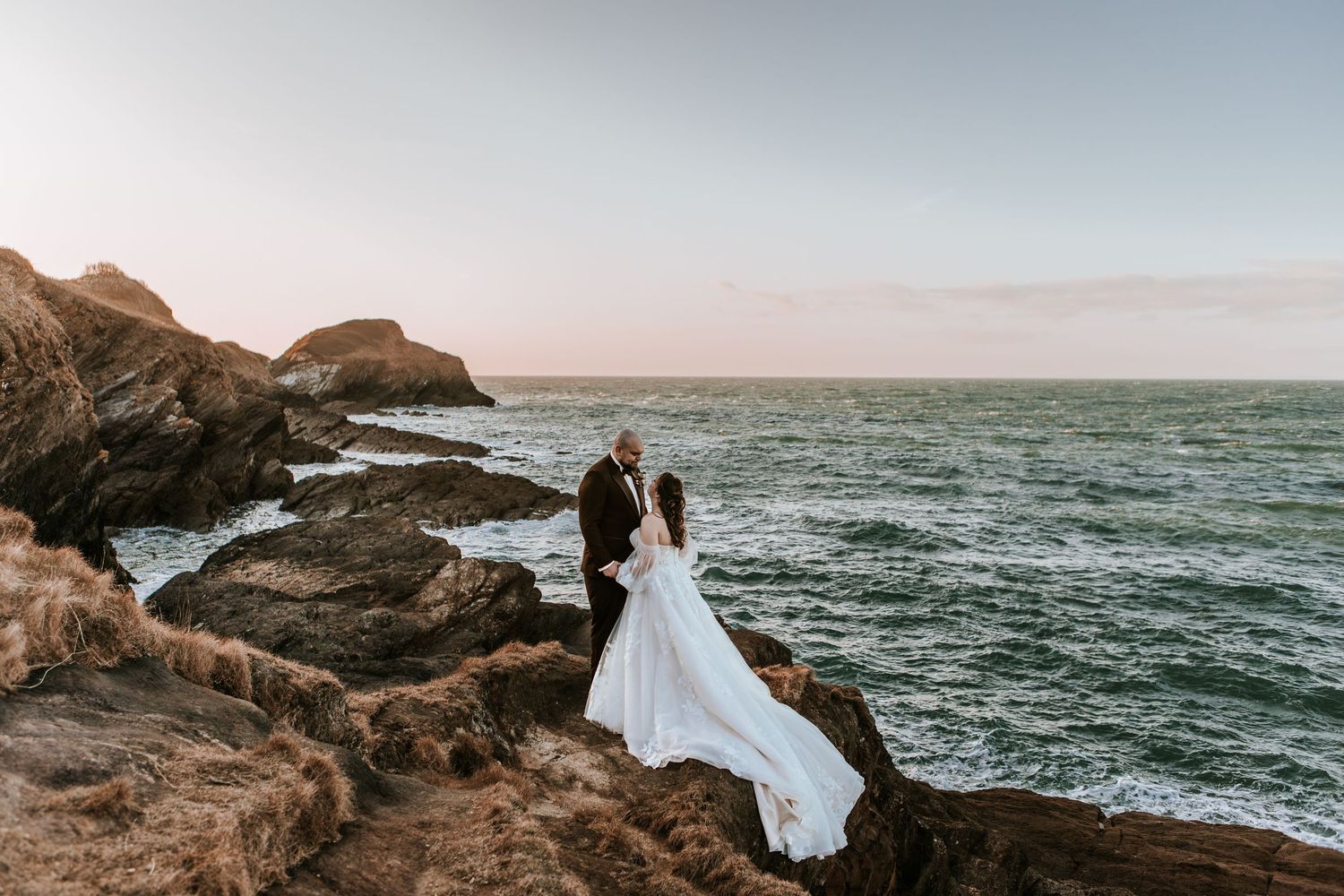 Bride and groom stand on rocky coastal cliffs overlooking ocean waves at sunset, wedding dress flowing in the wind.