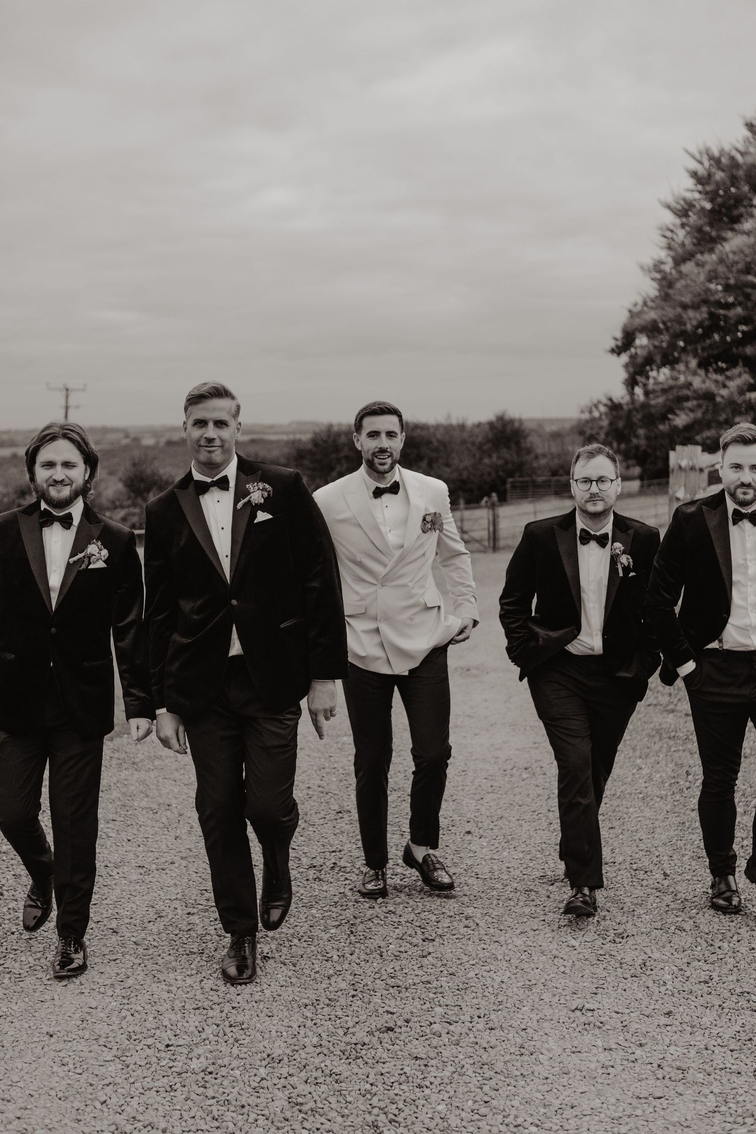 A group of groomsmen in black tuxedos walk together on a gravel path in black and white.