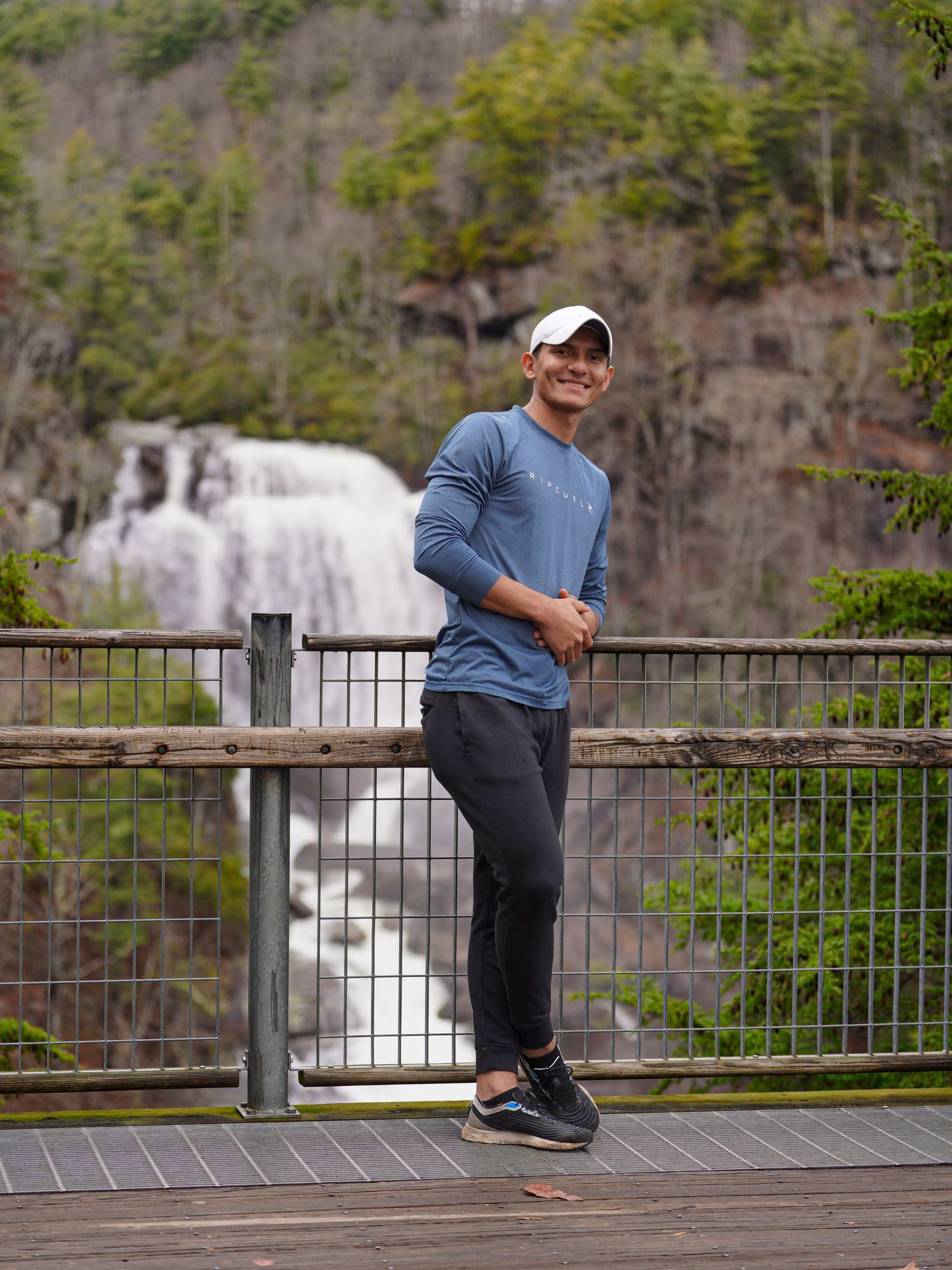 A person strikes playful poses on a wooden observation deck overlooking a scenic waterfall in the background.