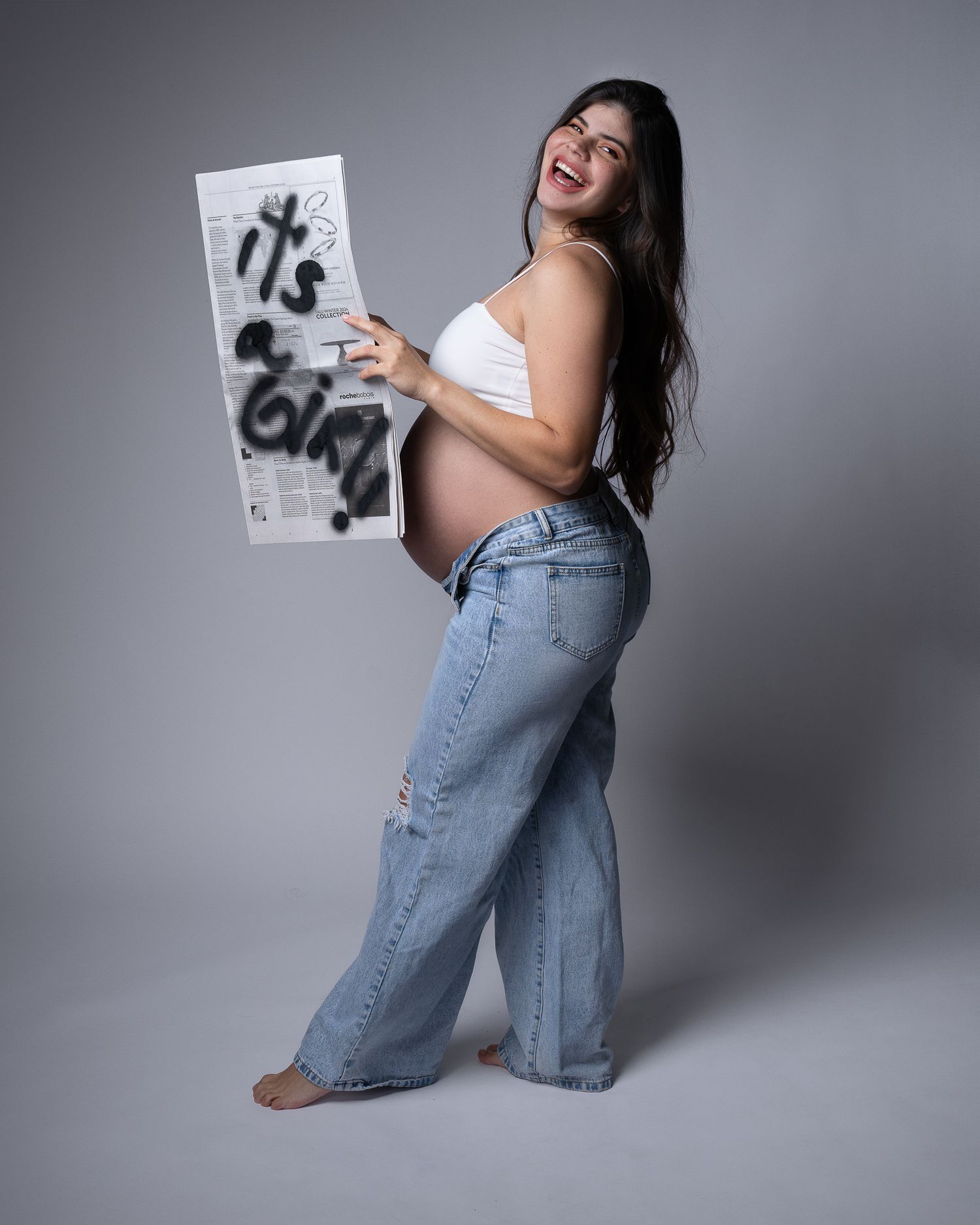 A person in light-colored top and jeans poses playfully against a gray studio backdrop while holding a magazine.