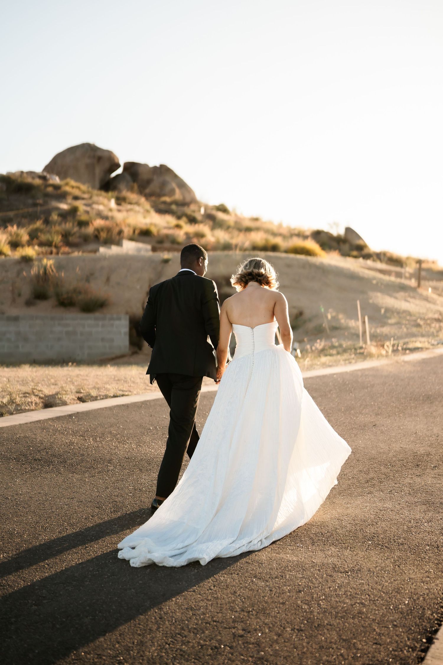 Newlyweds walking hand in hand along a desert path at sunset.