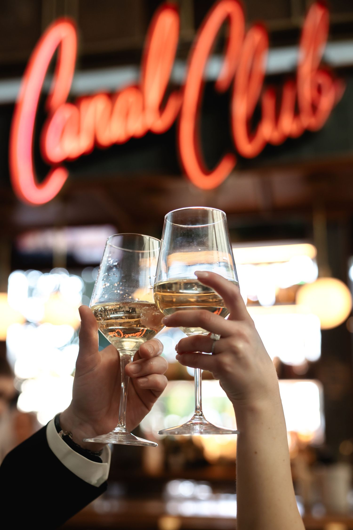 Two hands toasting wine glasses under red neon 'Canal Club' sign.