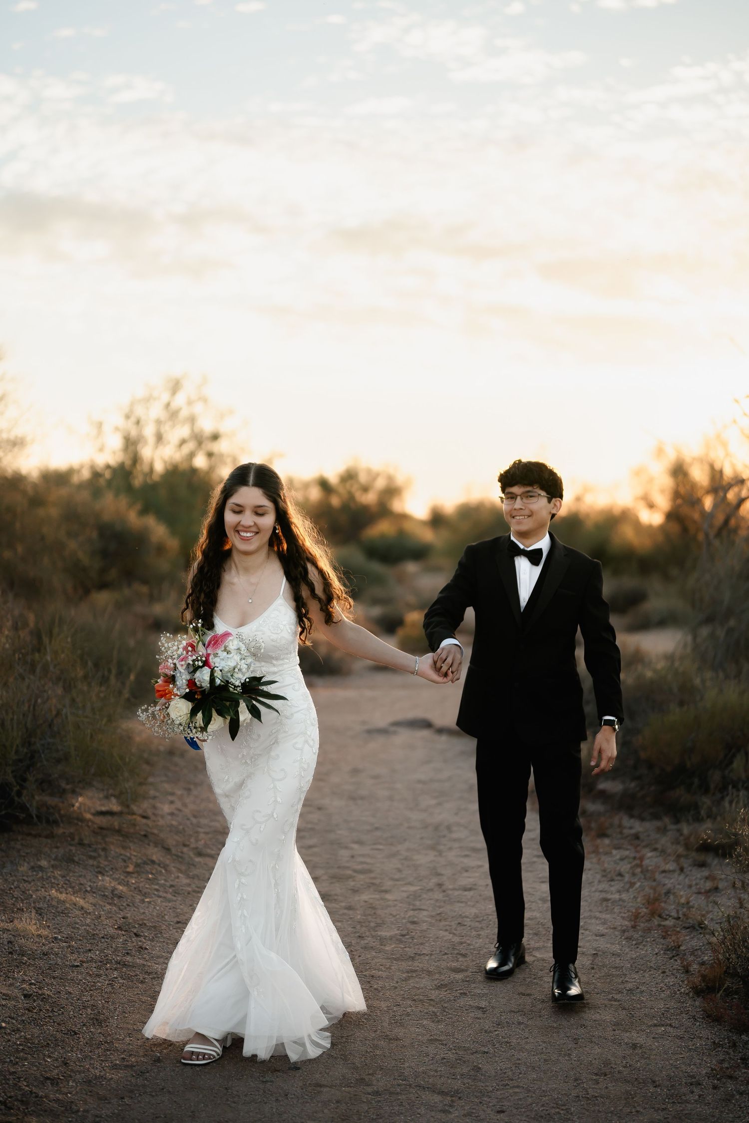 A couple walks hand in hand along a desert path at sunset, wearing formal wedding attire.