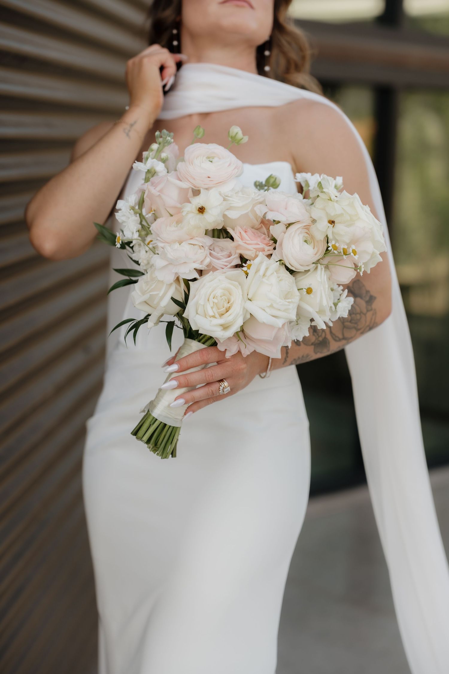 Bride in white halter gown holding elegant blush and ivory rose bouquet, showcasing tattoos and engagement ring.