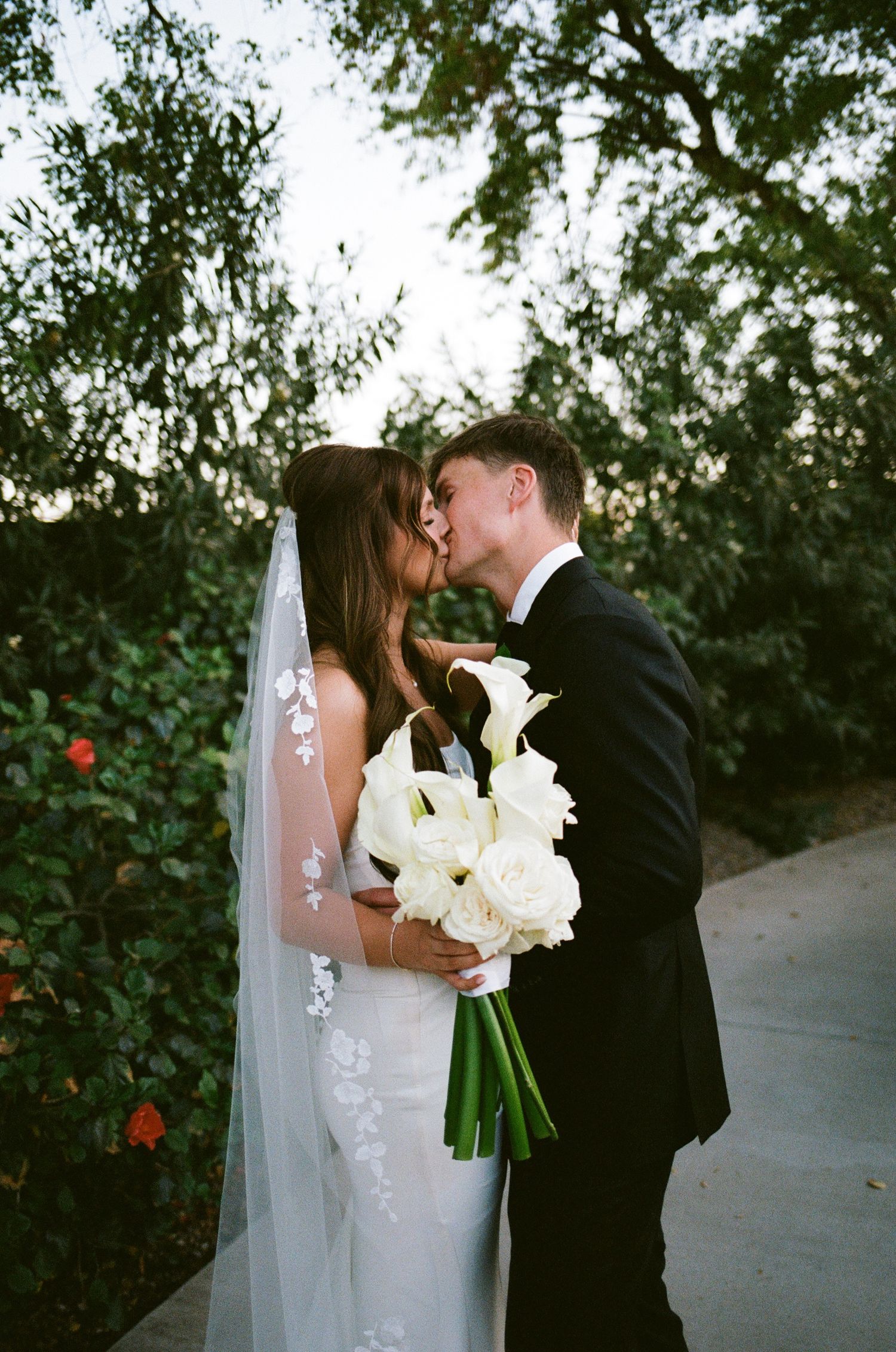 Bride and groom sharing a kiss outdoors, bride holding white calla lily bouquet and wearing a veil with floral embroidery.
