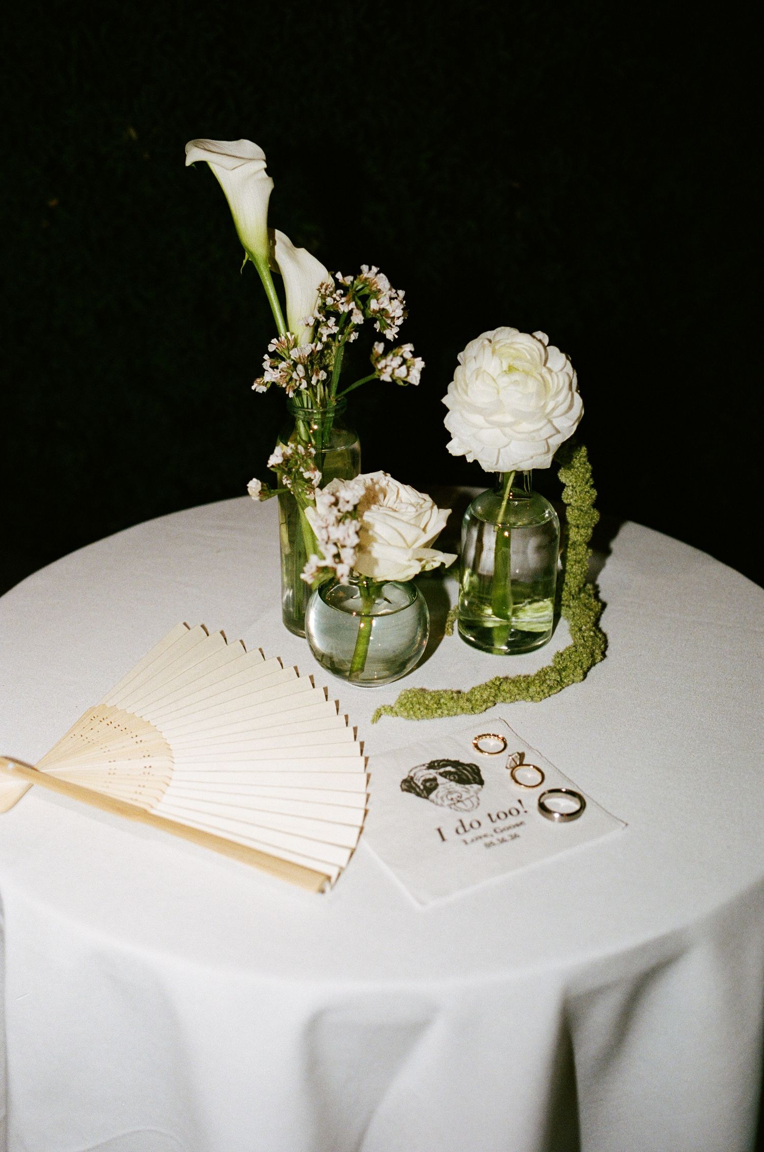 Elegant wedding table with white floral arrangements in glass vases, a decorative fan, rings, and an 