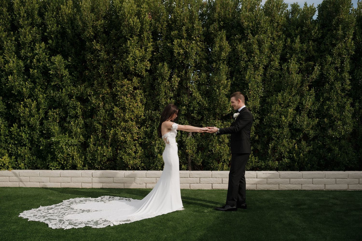 A couple in wedding attire holds hands while standing on a manicured lawn with a hedge backdrop.