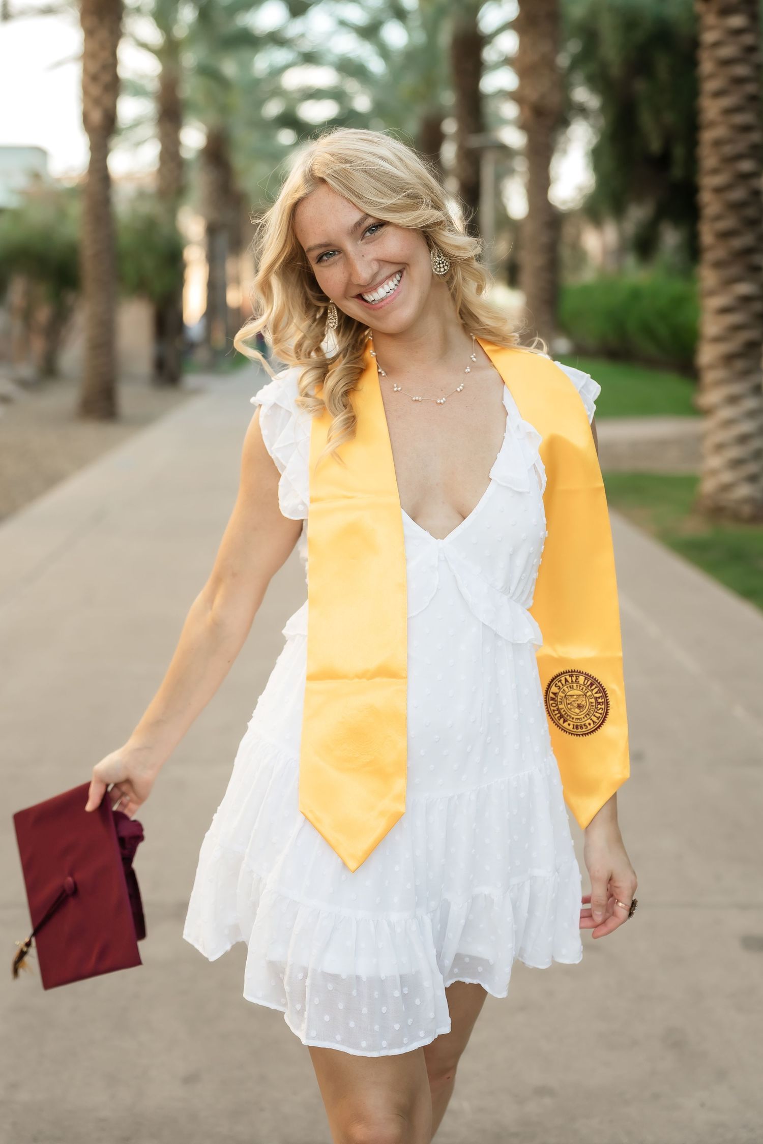 Graduate in white dress and yellow stole poses with diploma in palm tree-lined path.