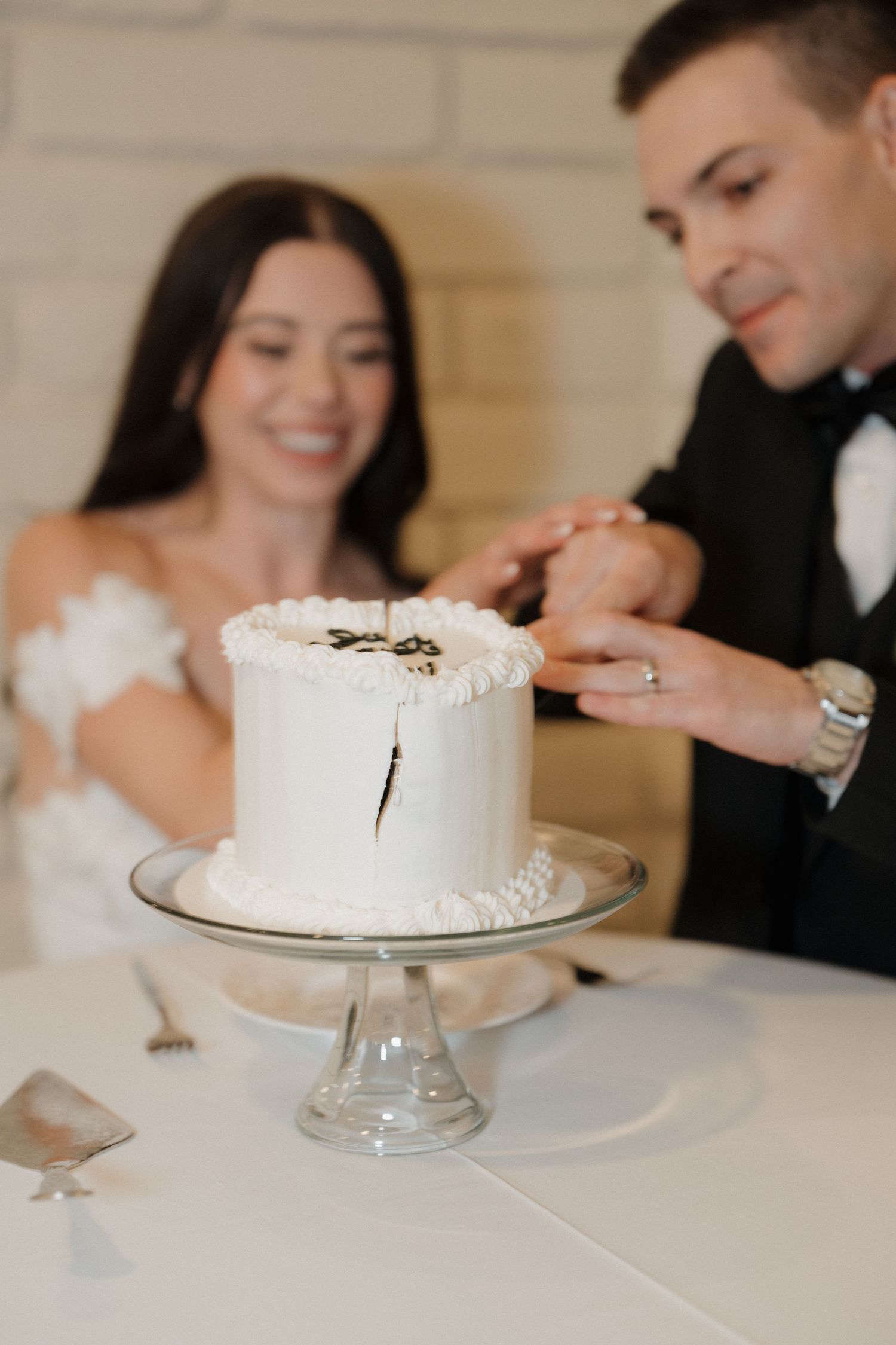 Elegant white wedding cake on a crystal cake stand being cut during a reception celebration.
