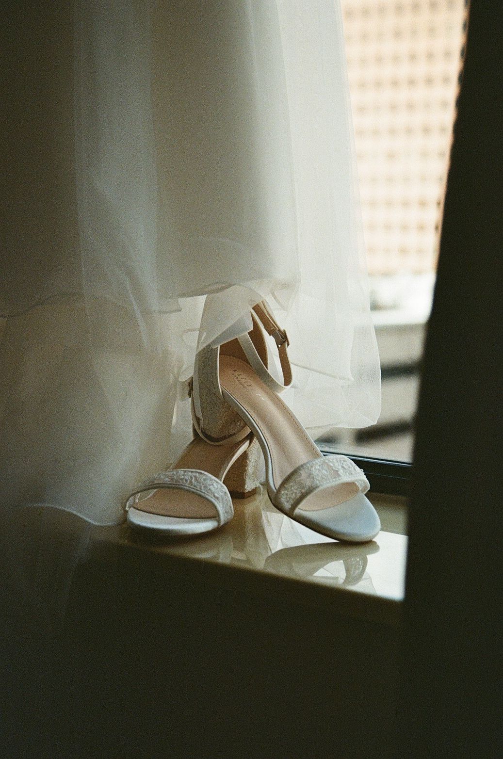 Elegant white bridal heels with ankle straps and block heels placed on a windowsill beside a flowing wedding veil.