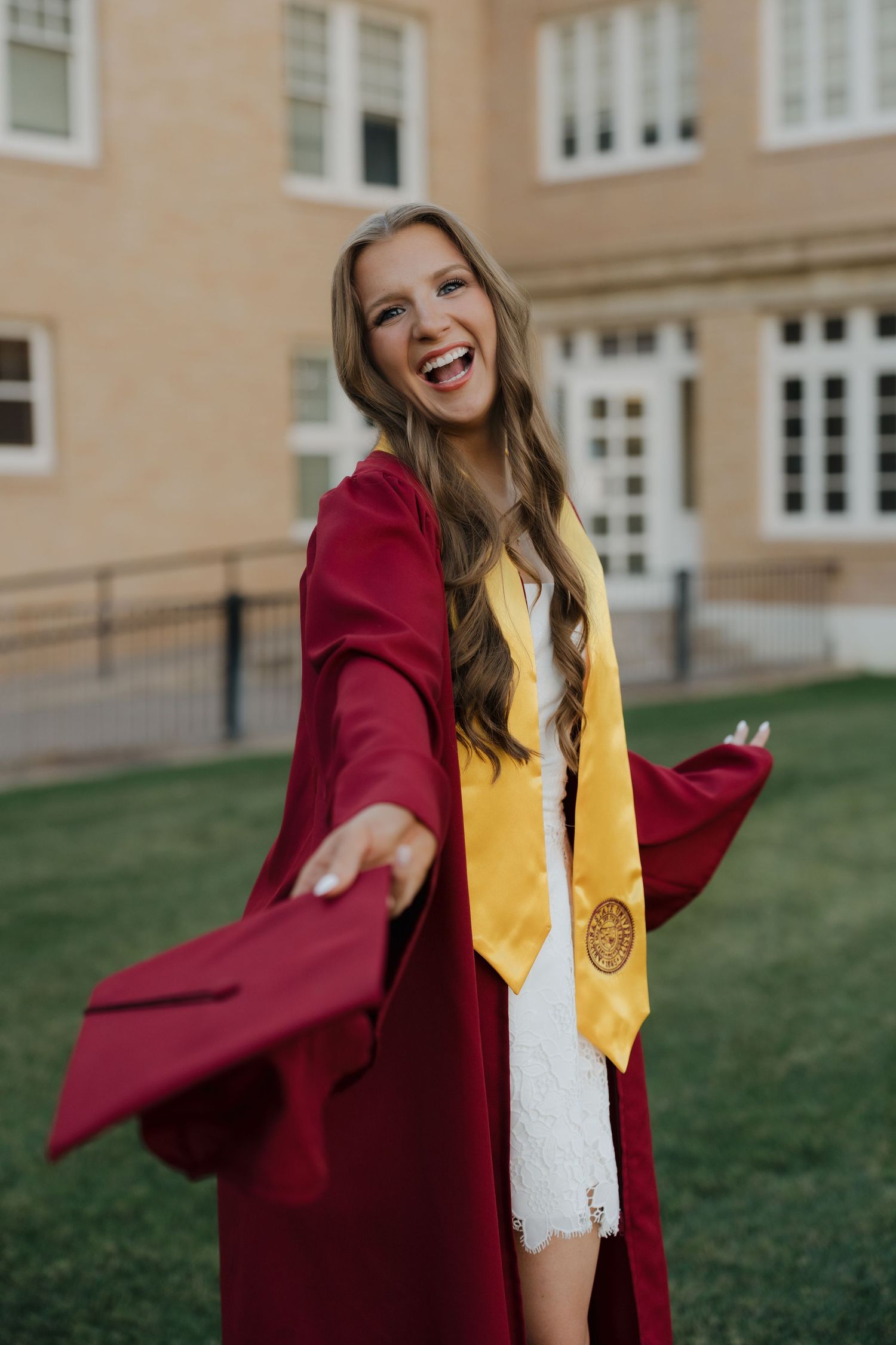 Smiling graduate in crimson gown and gold honor stole celebrates outdoors on campus lawn.