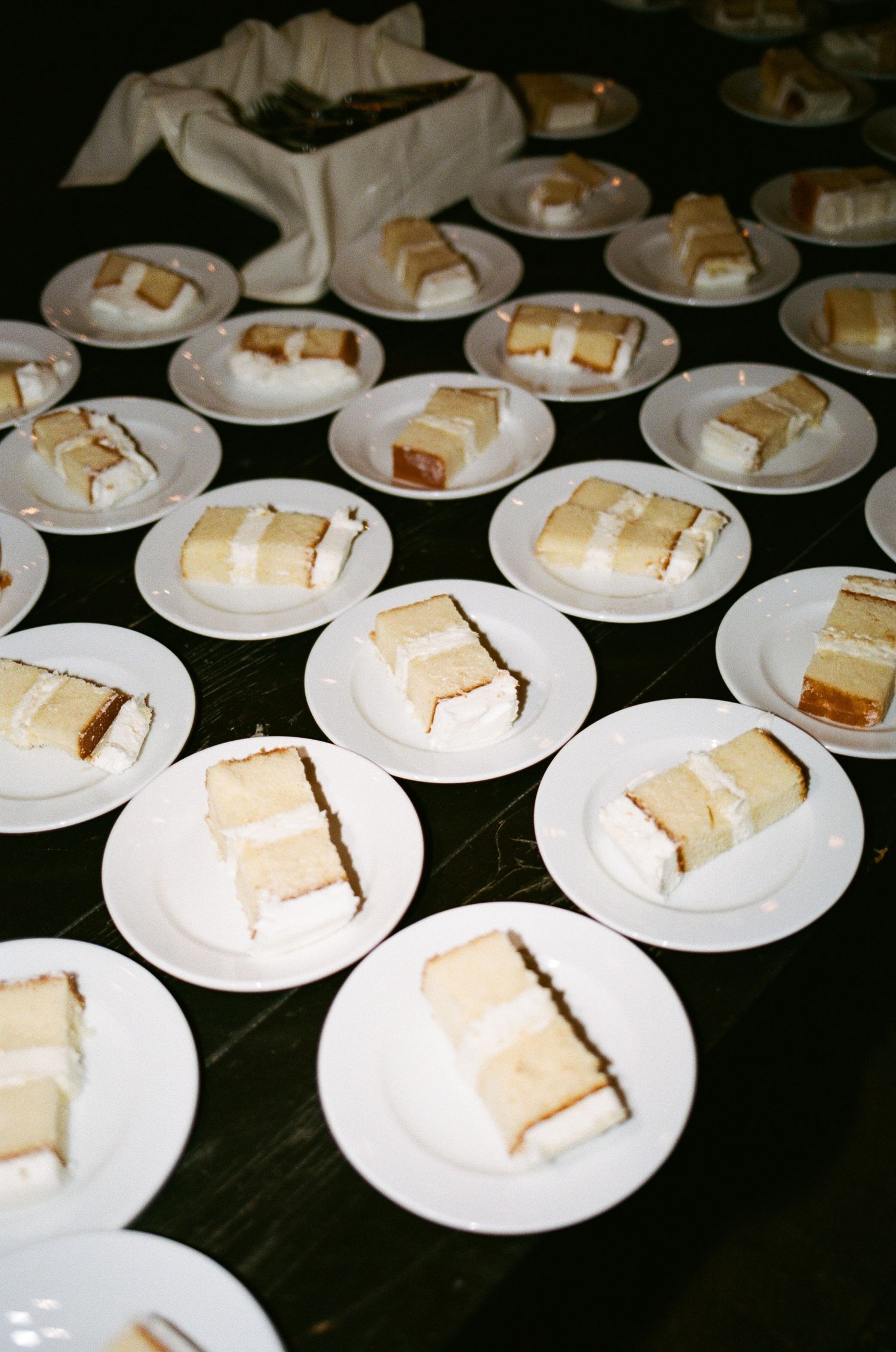 Multiple white plates of layered cake slices with white frosting arranged on a black tablecloth at a catered event.