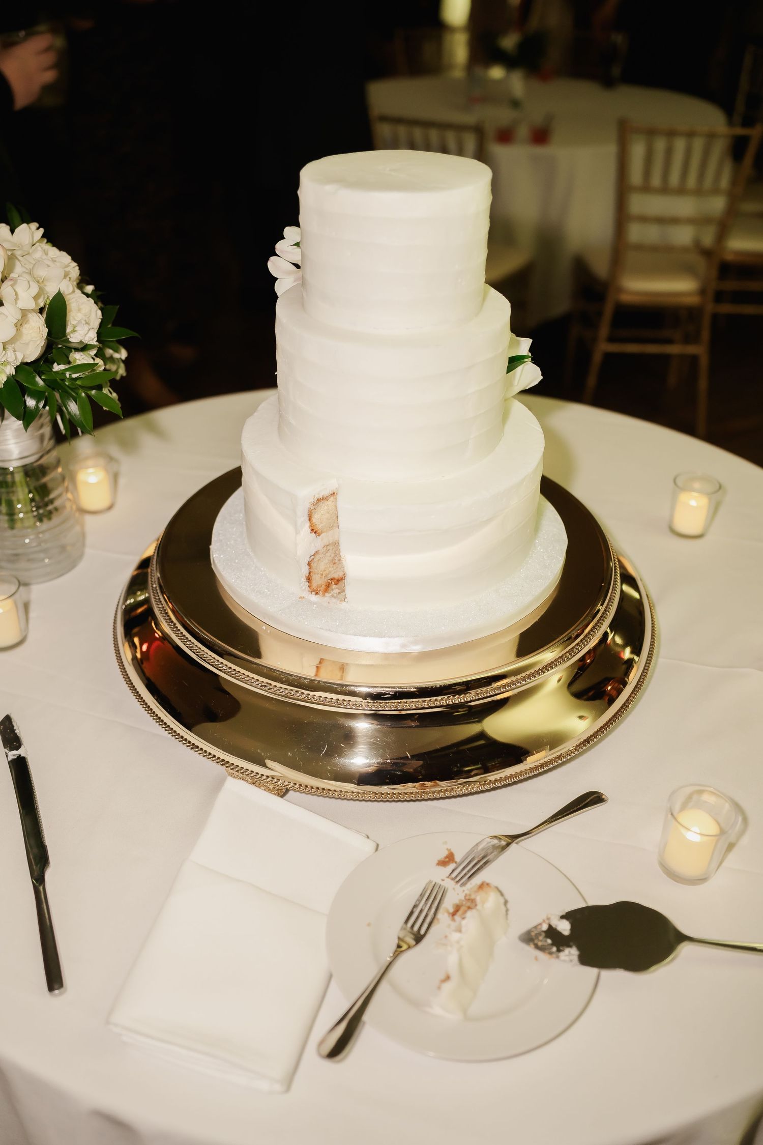 Simple white wedding cake displayed on silver platter with tea lights on reception table.
