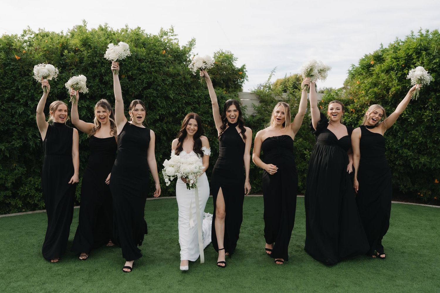 Wedding party in black dresses and white gown jumping joyfully on green lawn during outdoor celebration.