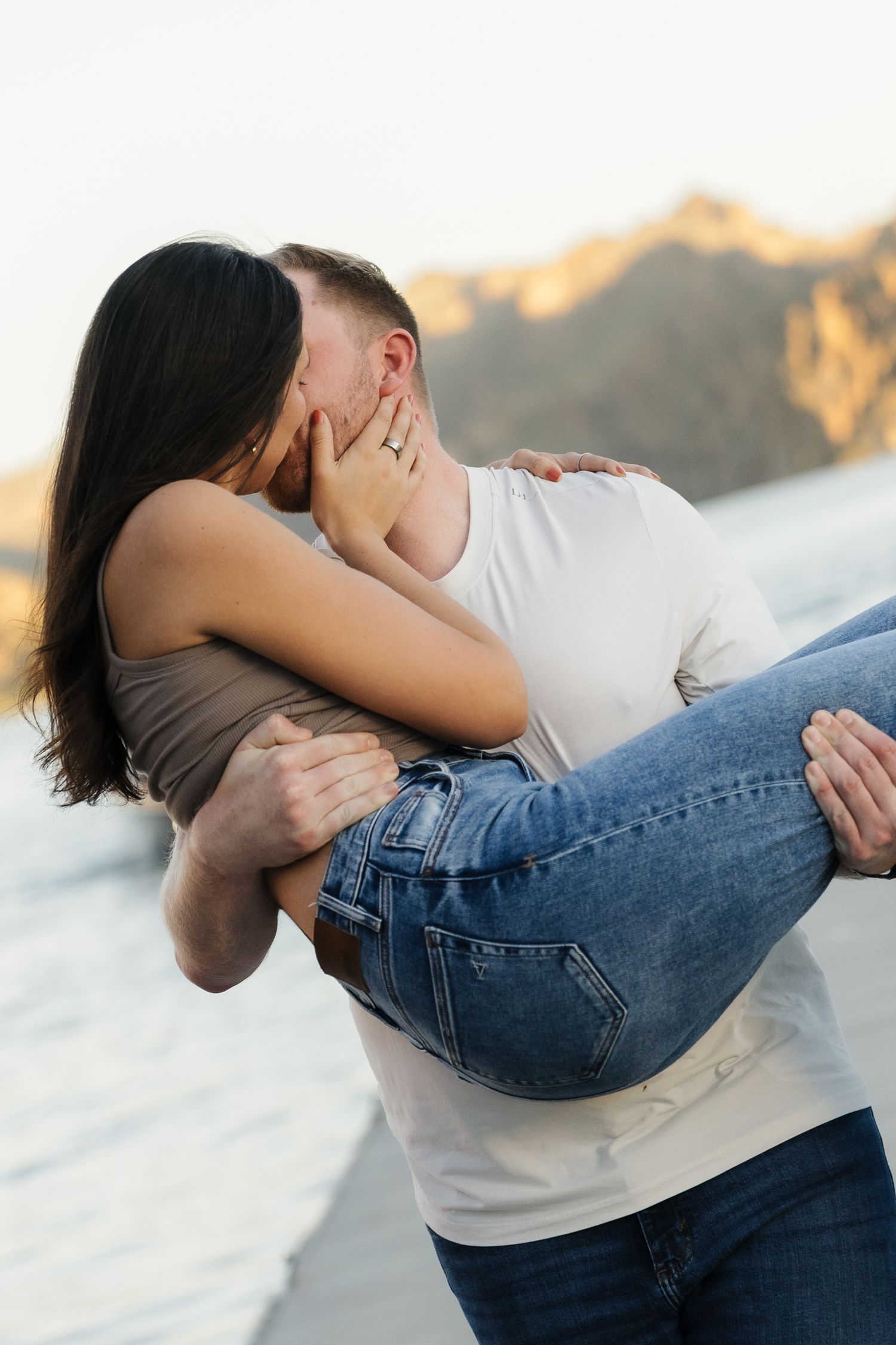 A romantic couple shares a passionate kiss on a beach at sunset, with the man carrying the woman in his arms.