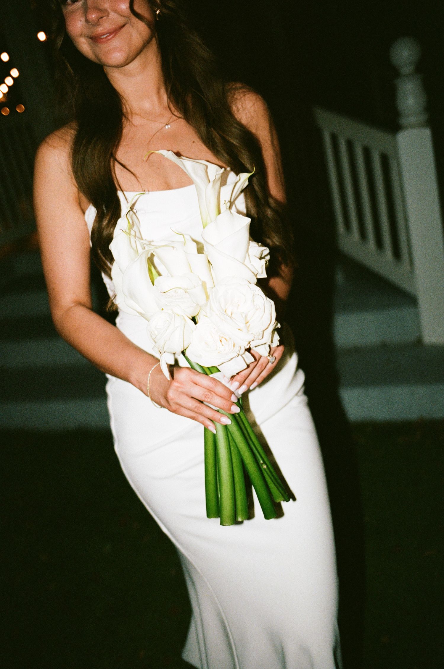 Bride in white strapless gown holding elegant white calla lily bouquet with green stems at outdoor evening wedding.
