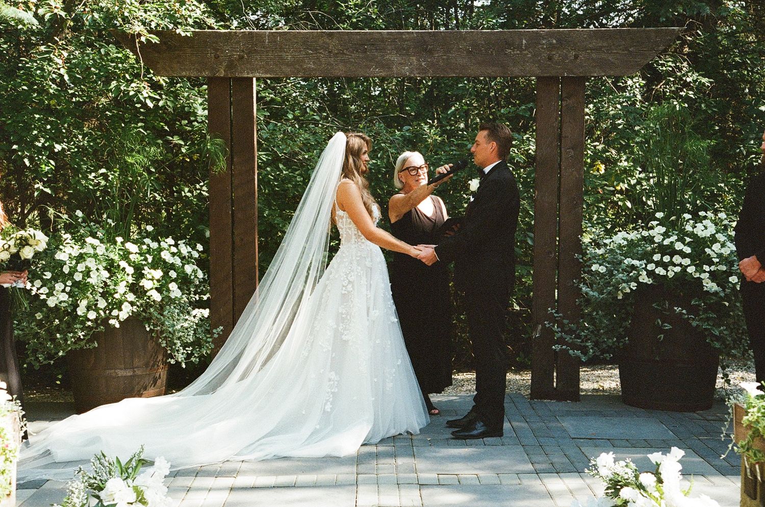 Bride and groom exchange vows under rustic wooden arch surrounded by lush greenery at outdoor garden wedding ceremony.