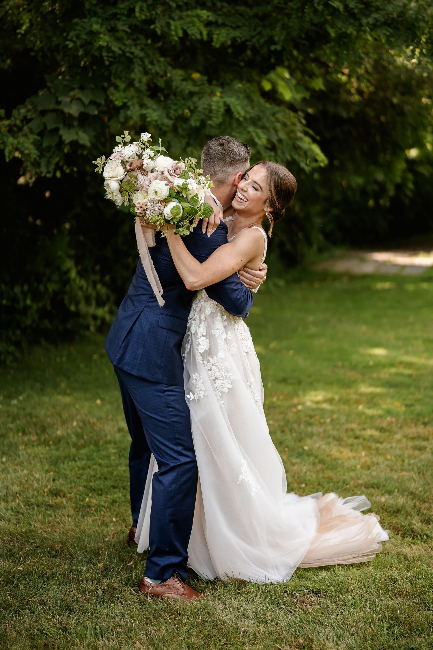 A couple in wedding attire shares a playful moment on grass with white floral bouquet.
