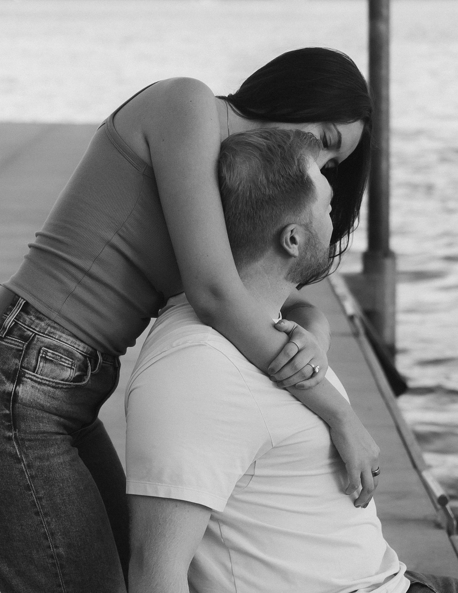 Black and white photo of a woman leaning over a seated man in an intimate embrace on a dock by the water.