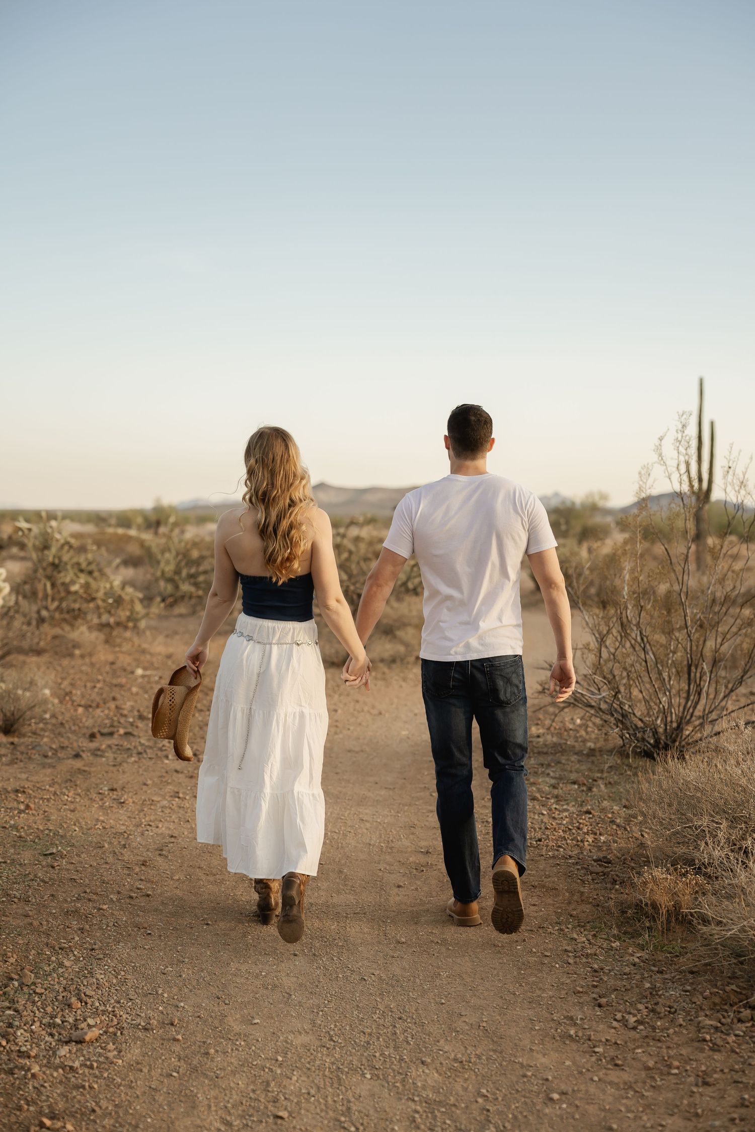 Couple walks together on desert trail at sunset with cacti in background.