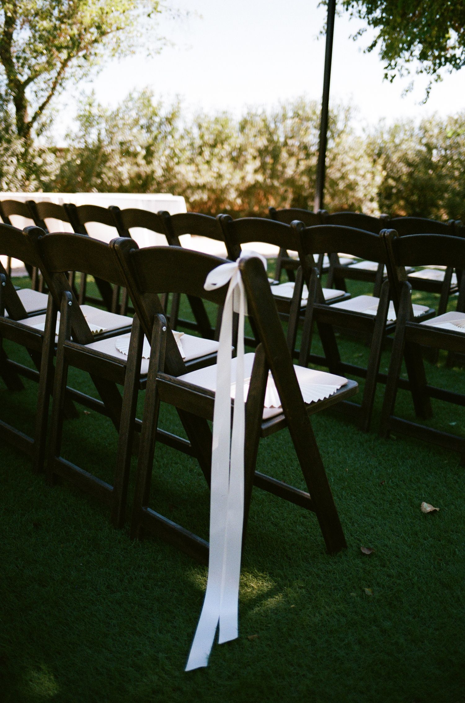 Dark wooden folding chairs with white ribbon bows arranged on green grass for an elegant outdoor wedding ceremony.