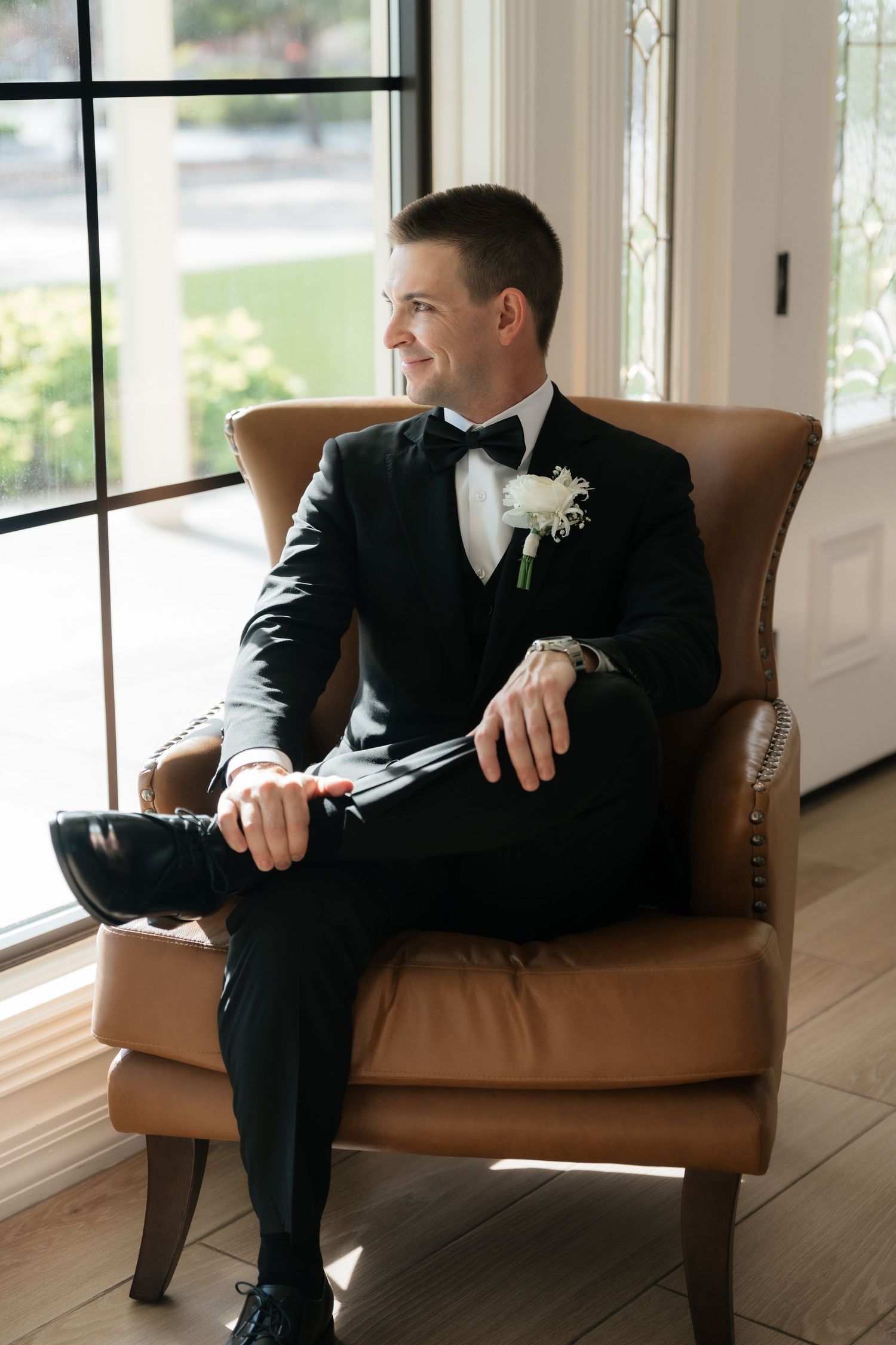 Groom in black tuxedo sitting on brown leather couch in sunlit room with hardwood floors.