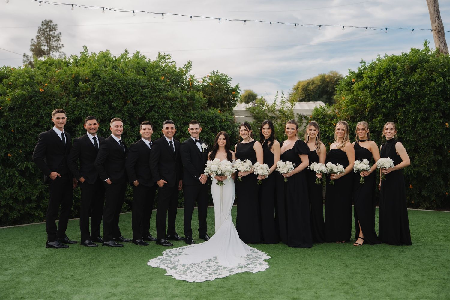 Wedding party in black attire poses outdoors in a garden setting with a flowing white wedding dress.