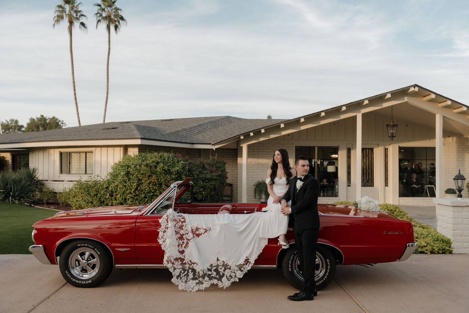 Classic red convertible car parked outside a venue with wedding party posing alongside it.