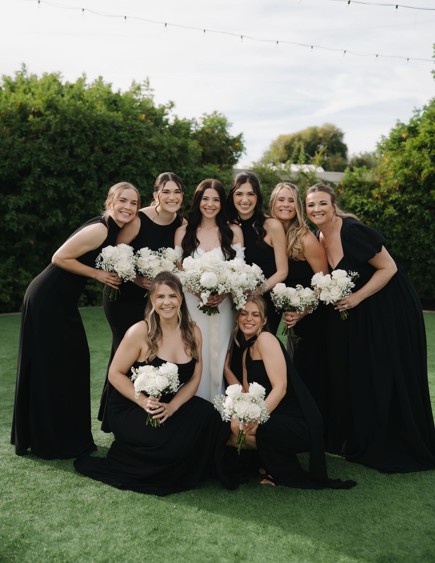 Wedding party in black dresses holding white bouquets poses together on green lawn.
