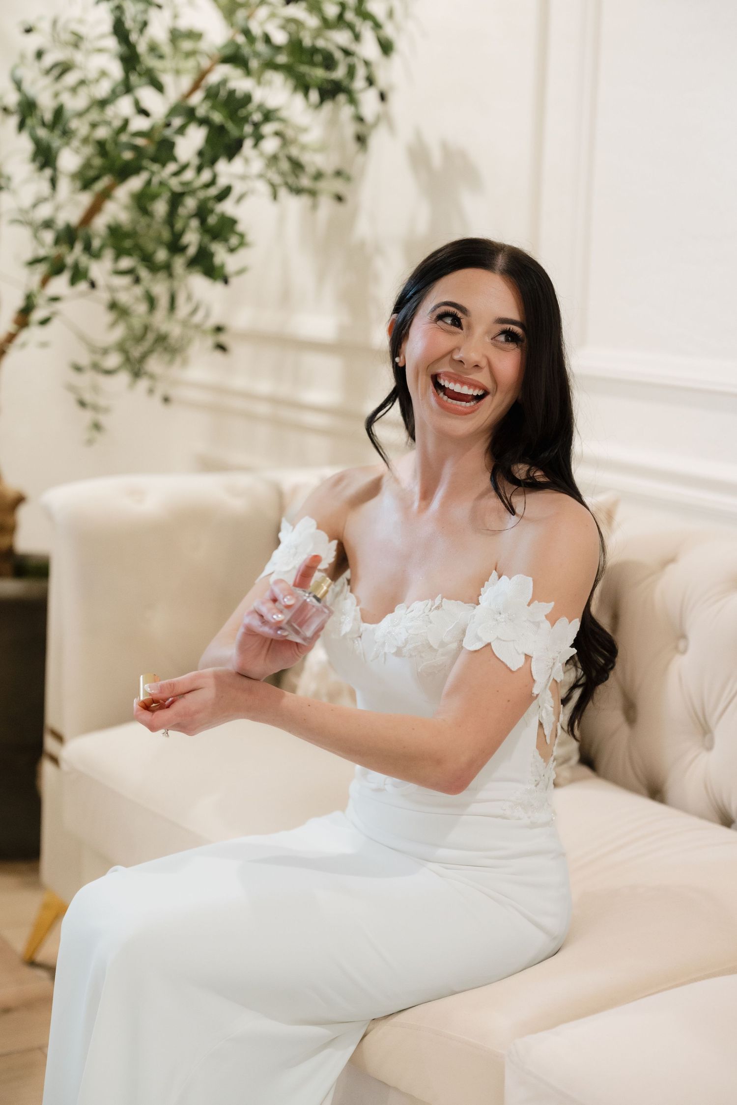 Joyful bride in white strapless gown with floral details laughs while sitting on cream colored couch beneath greenery.