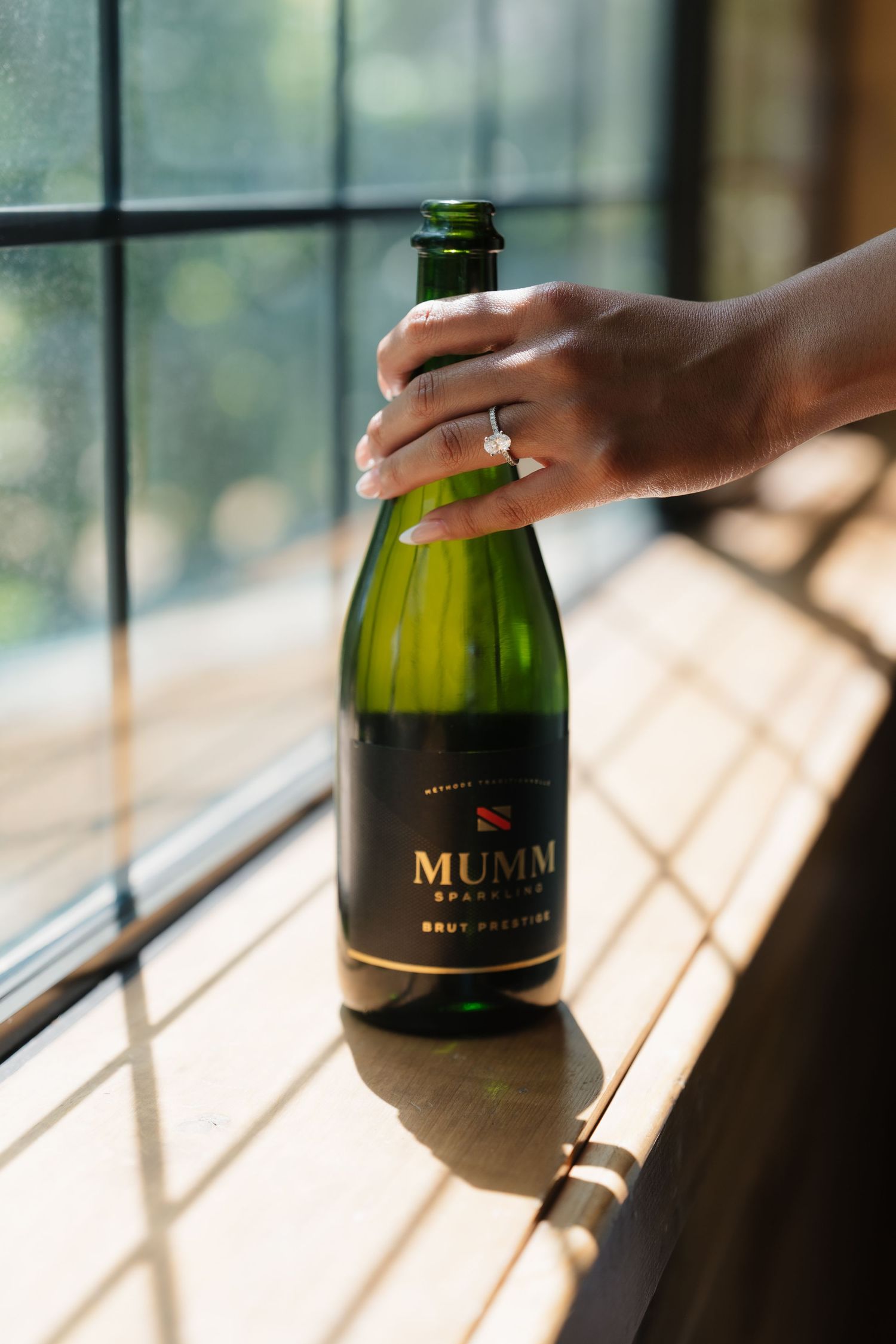 Hand holding a Mumm Sparkling Brut Prestige champagne bottle on a wooden ledge by a sunlit window.