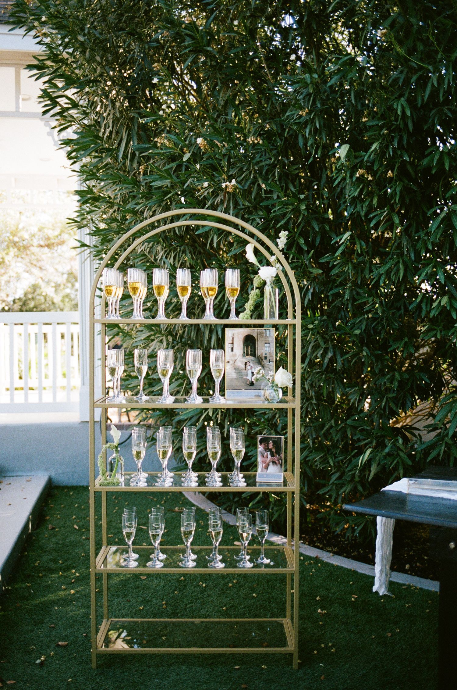Gold arched display shelf with champagne flutes arranged on multiple tiers, set outdoors against lush green foliage.